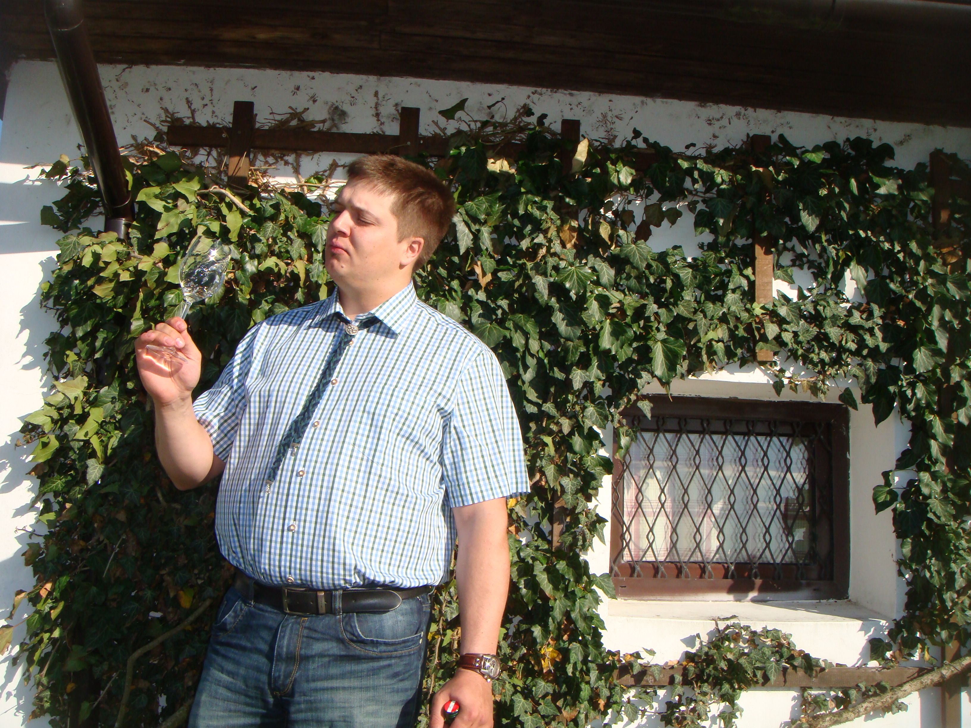 Man in plaid shirt looks at a wine glass in front of a wall covered with ivy.