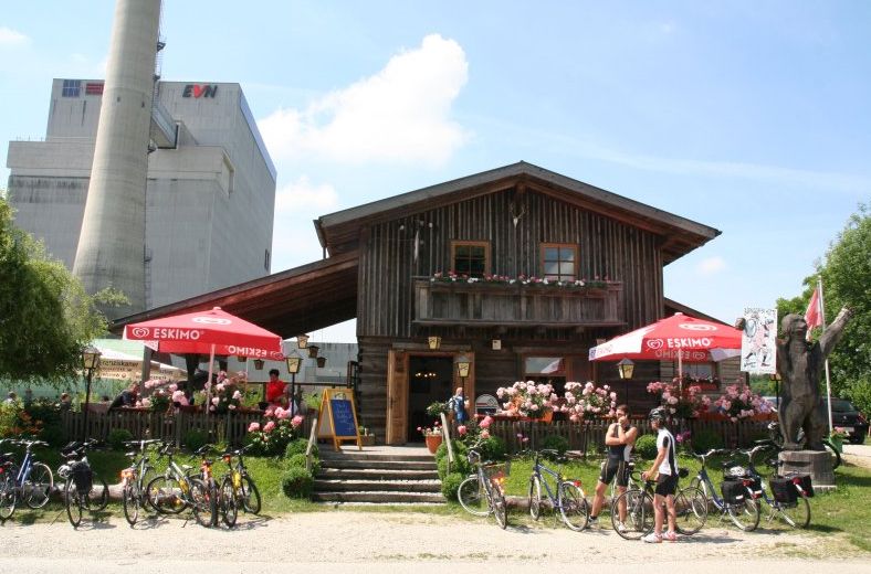 Wooden hut with terrace, bicycles and parasols in front of an industrial building.
