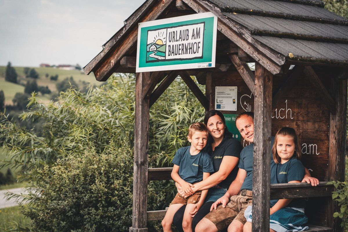 The Korntheuer family is sitting in a wooden shelter with a sign that reads "Farm Vacation." Green hills can be seen in the background.