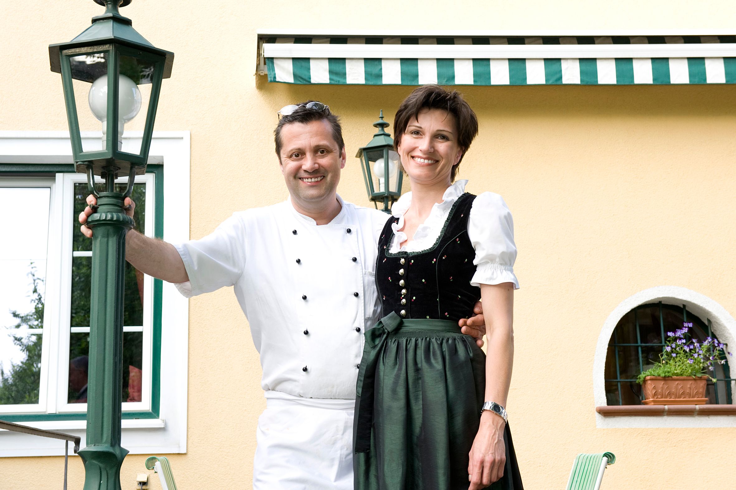 A man in chef's clothes and a woman in traditional costume stand smiling in front of a yellow building with green lanterns.