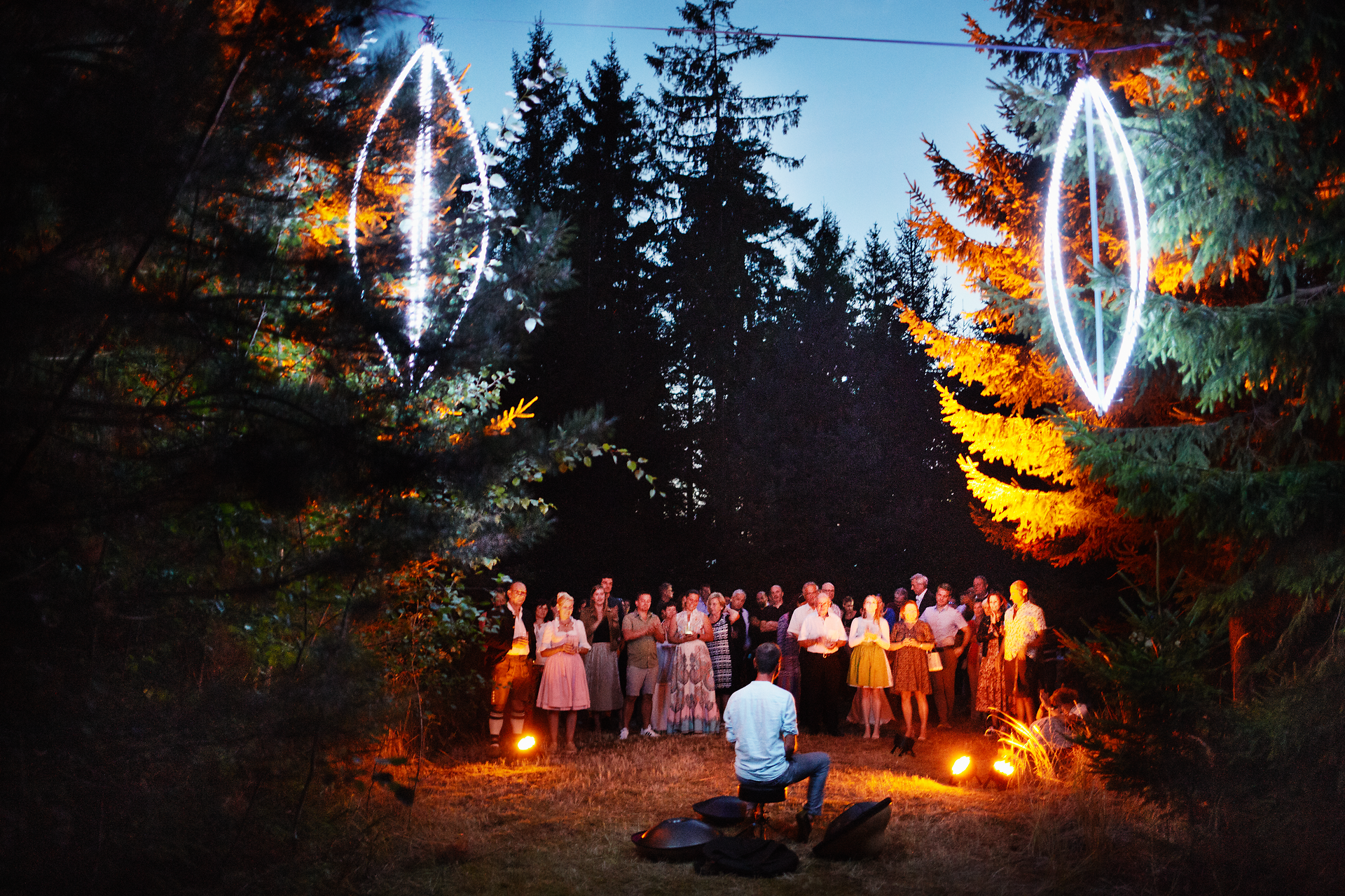 Inmitten eines malerischen Waldes versammeln sich Menschen, um die harmonischen Klänge eines Musikers zu genießen. Die sanften Lichter der Umgebung schaffen eine zauberhafte Atmosphäre, die den Sommerabend unvergesslich macht.