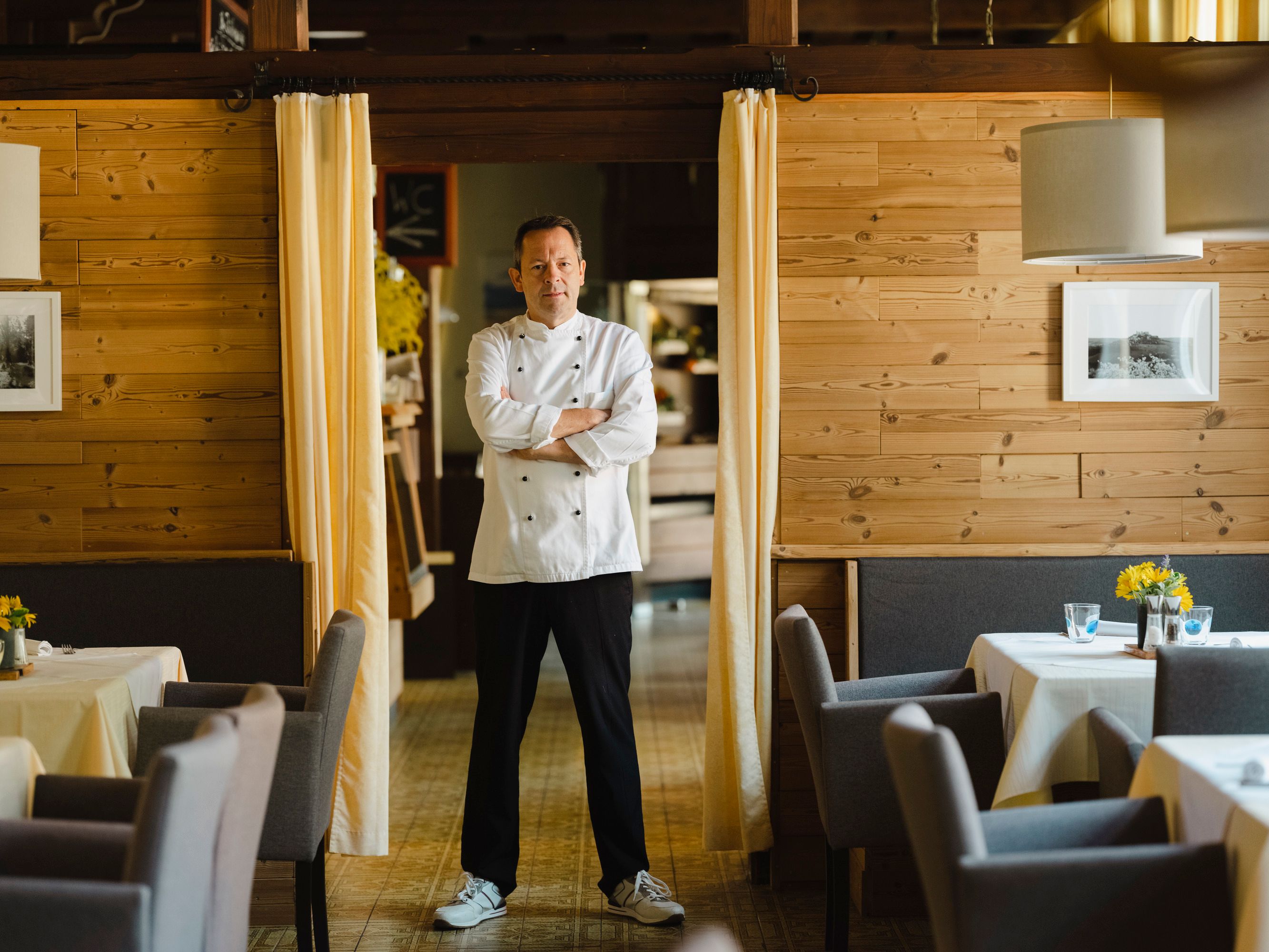 A man in chef's clothes stands with his arms folded in a restaurant with wooden walls.