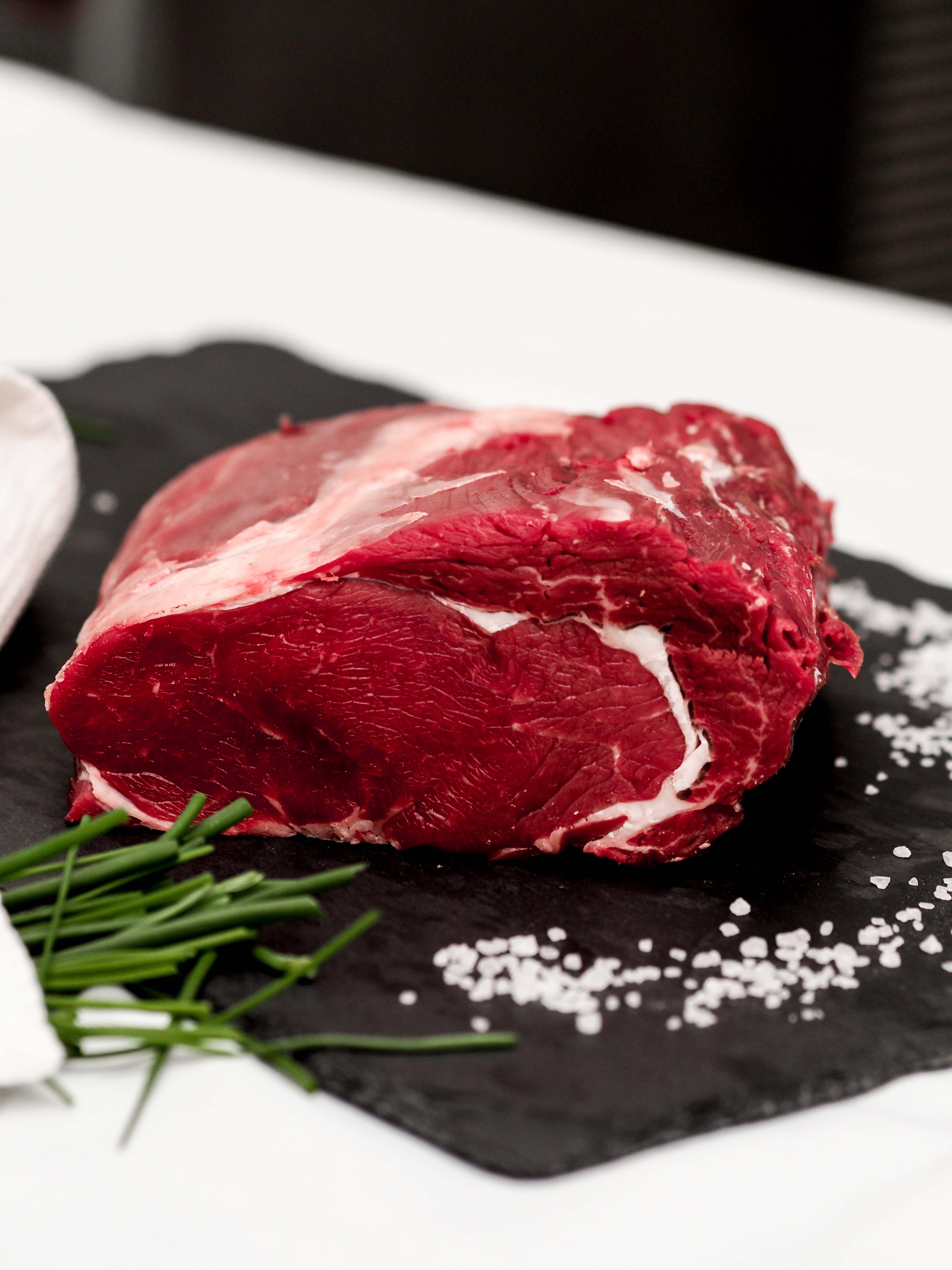 A raw piece of beef lies on a dark slate next to salt and chives.