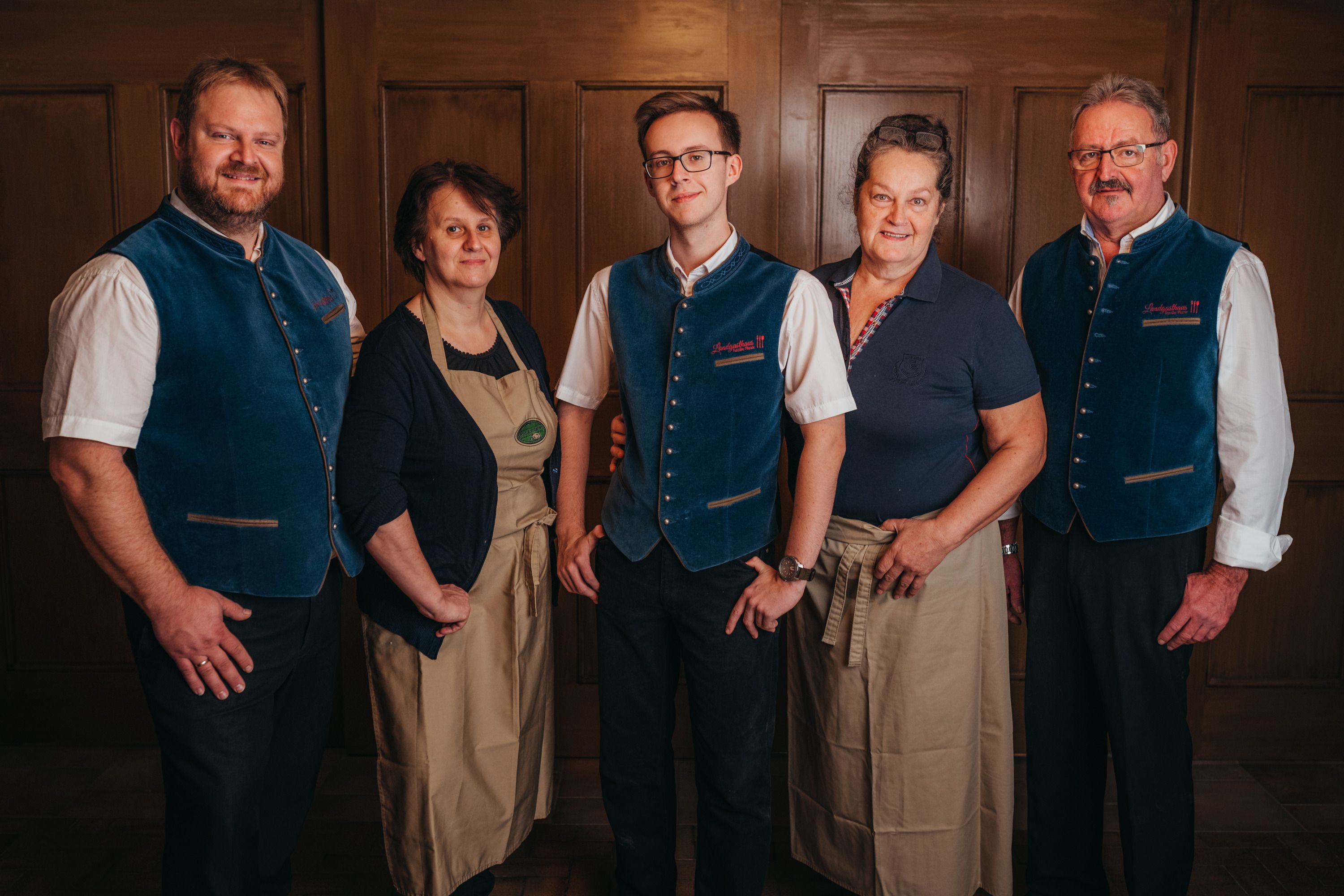 Five people in traditional dress stand in front of a wooden wall.
