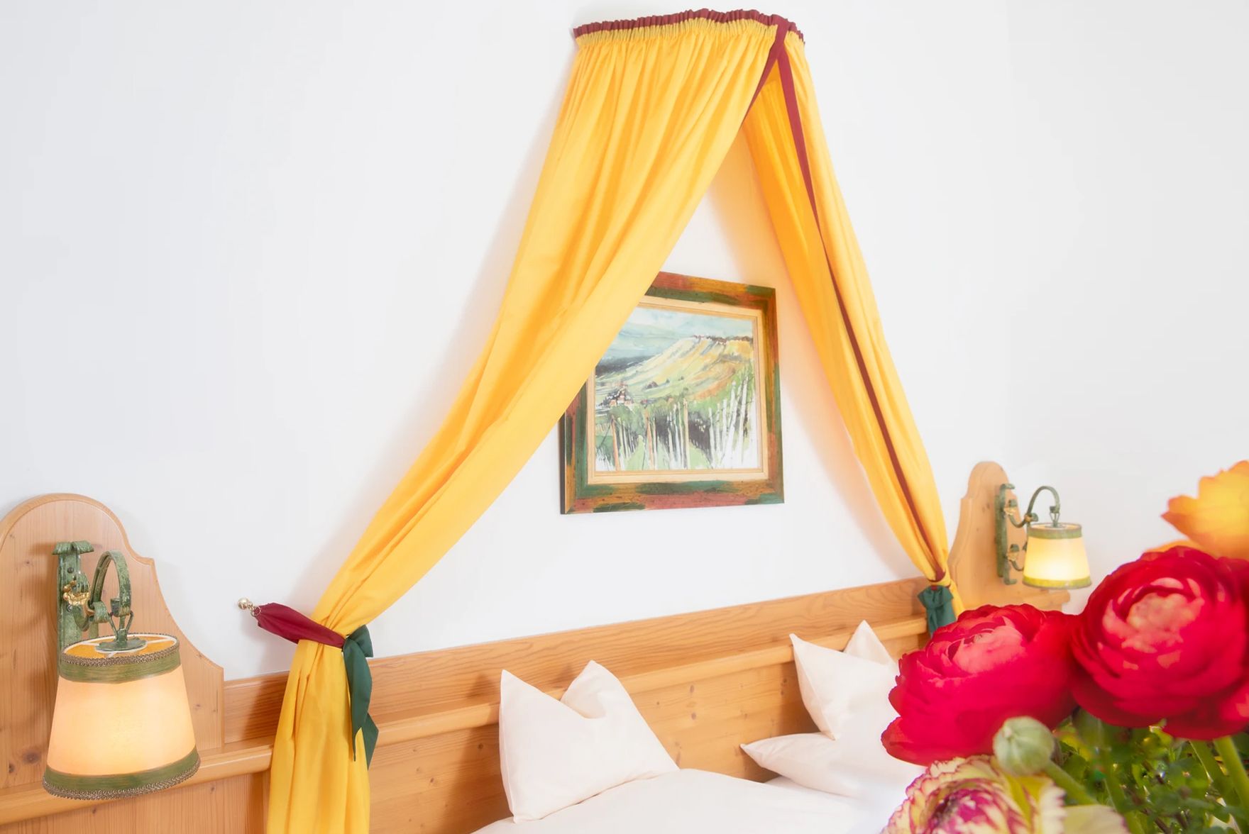 A cozy double room with a yellow canopy, wooden bed, wall lamps and a bouquet of flowers in the foreground.