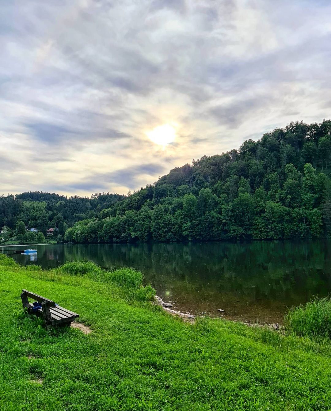 A bench stands on a green meadow on the banks of the Thurnberg reservoir, surrounded by wooded hills and under a cloudy sky with visible sun.