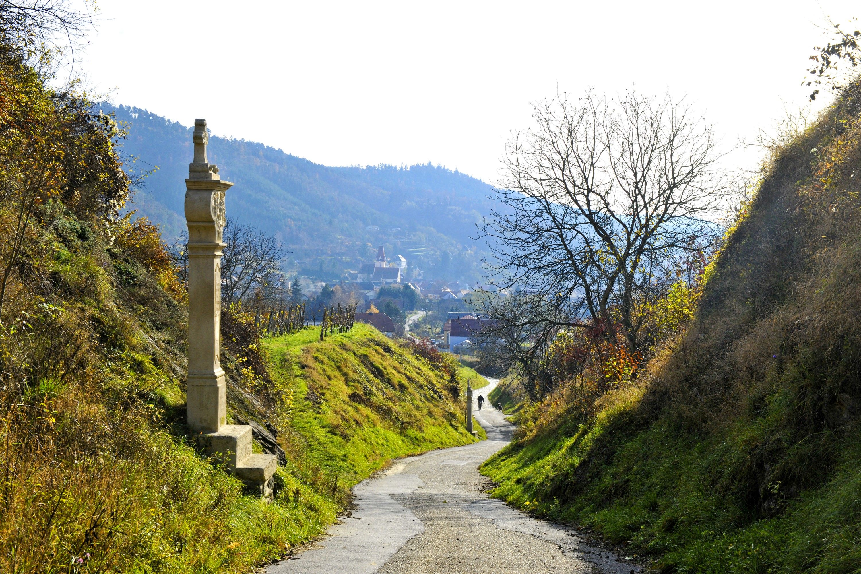 A narrow path leads through a hilly landscape with a stone monument and bare trees in the Kamp Valley ("Kamptal" in German) Schönberg Nature Park.