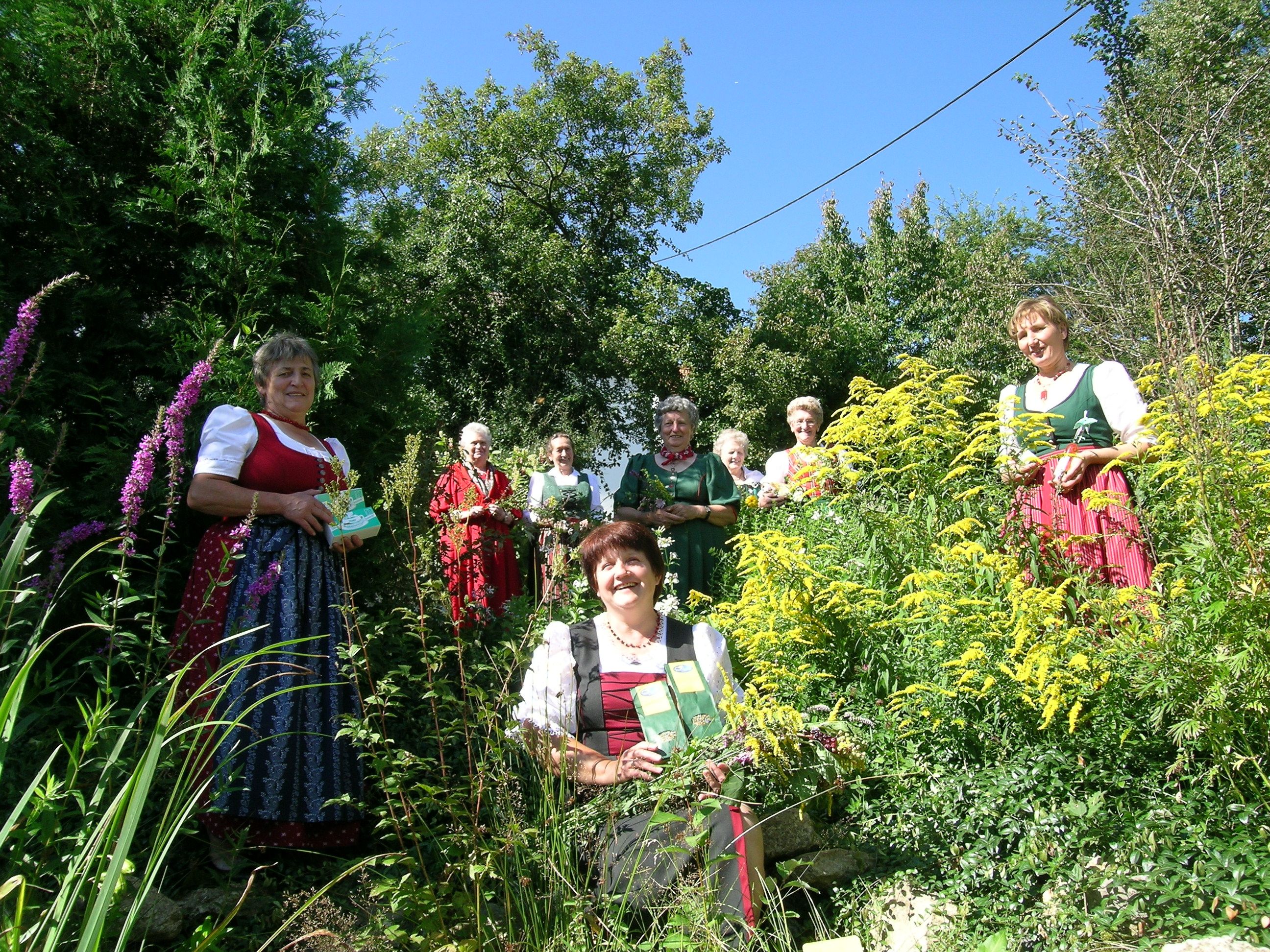 Women in traditional dress stand in a garden with herbs and flowers.