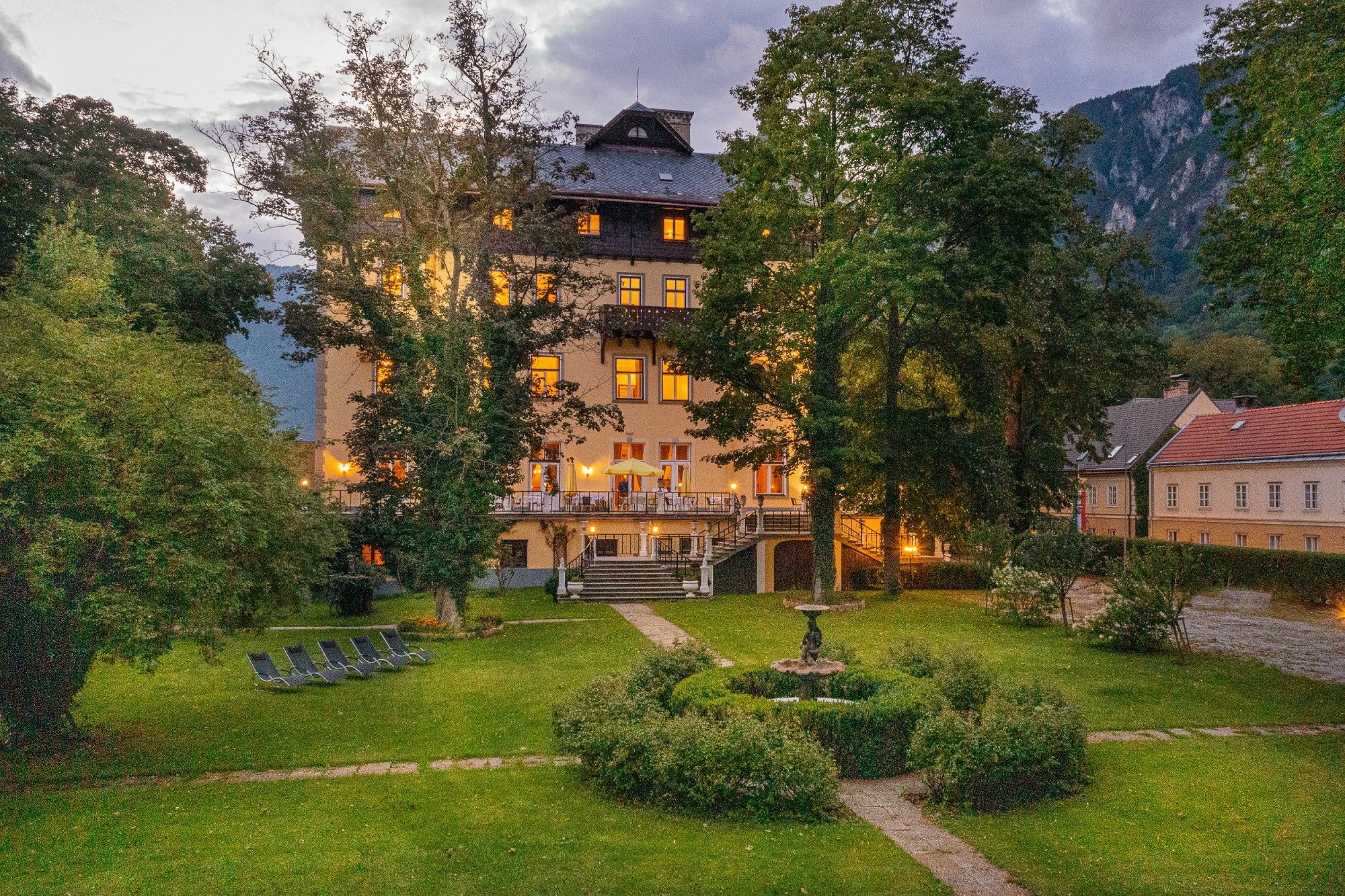 Illuminated building in a garden at dusk, surrounded by trees and mountains.