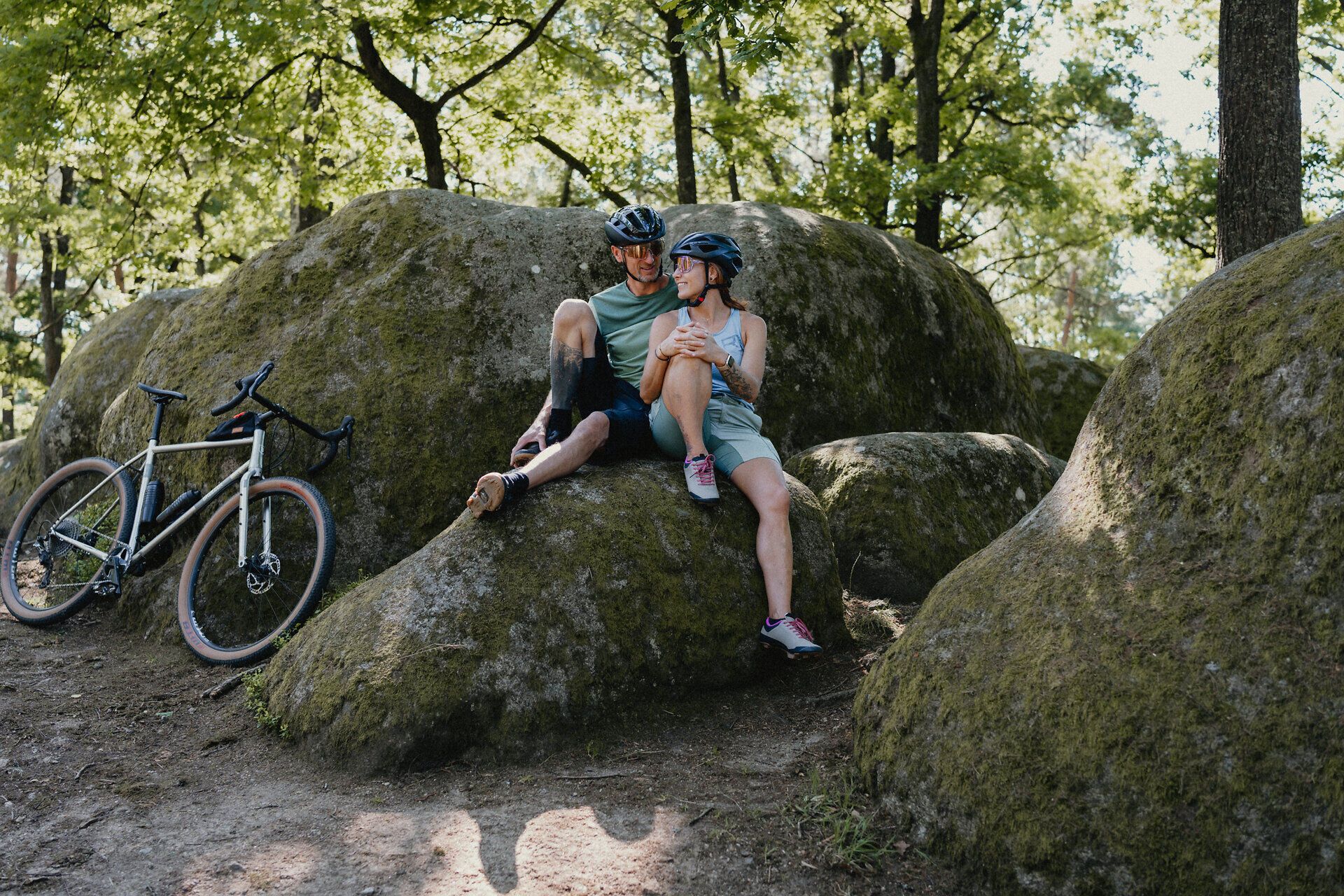 A man and a woman in bicycle gear sit on a stone and talk, their bicycles leaning on another stone next to them