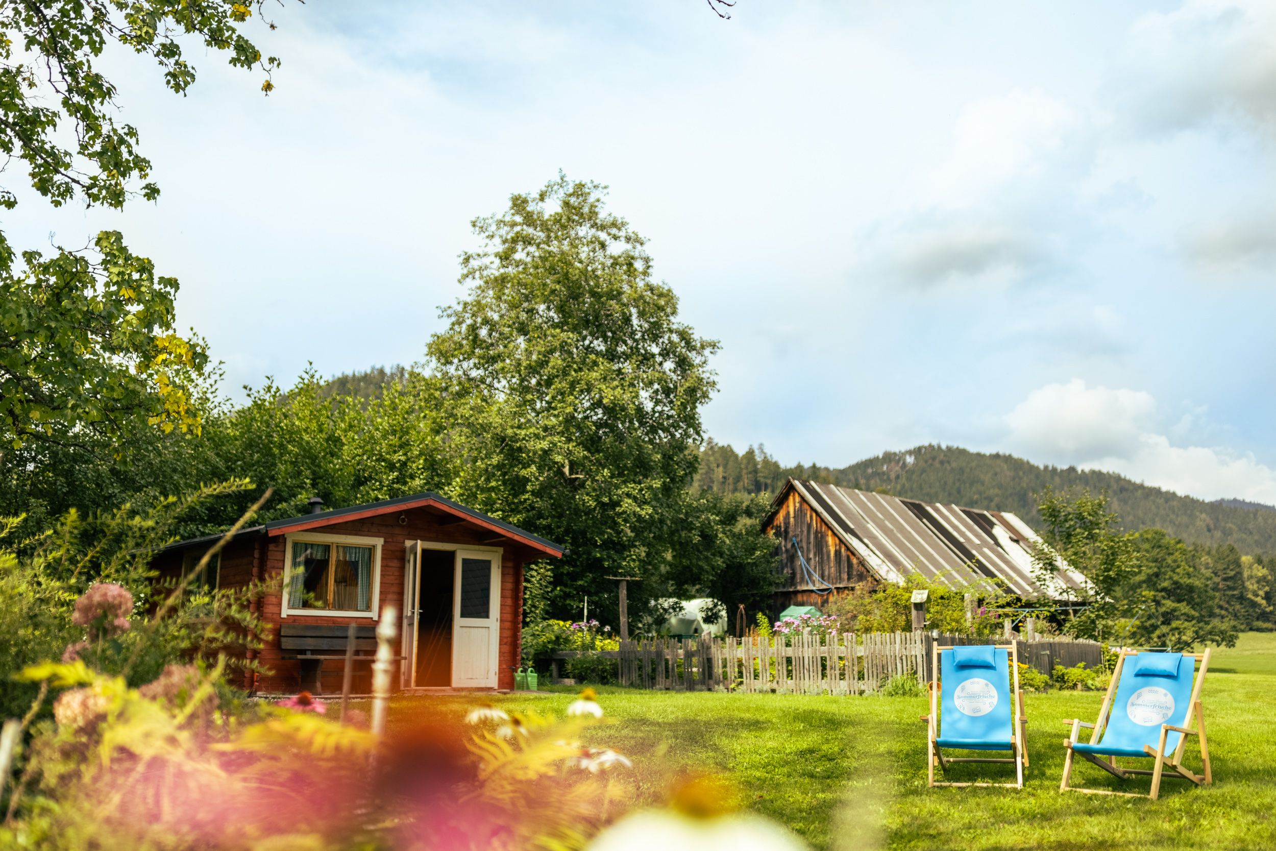 An idyllic garden with a small wooden house, two blue deckchairs and an old barn building in the background.