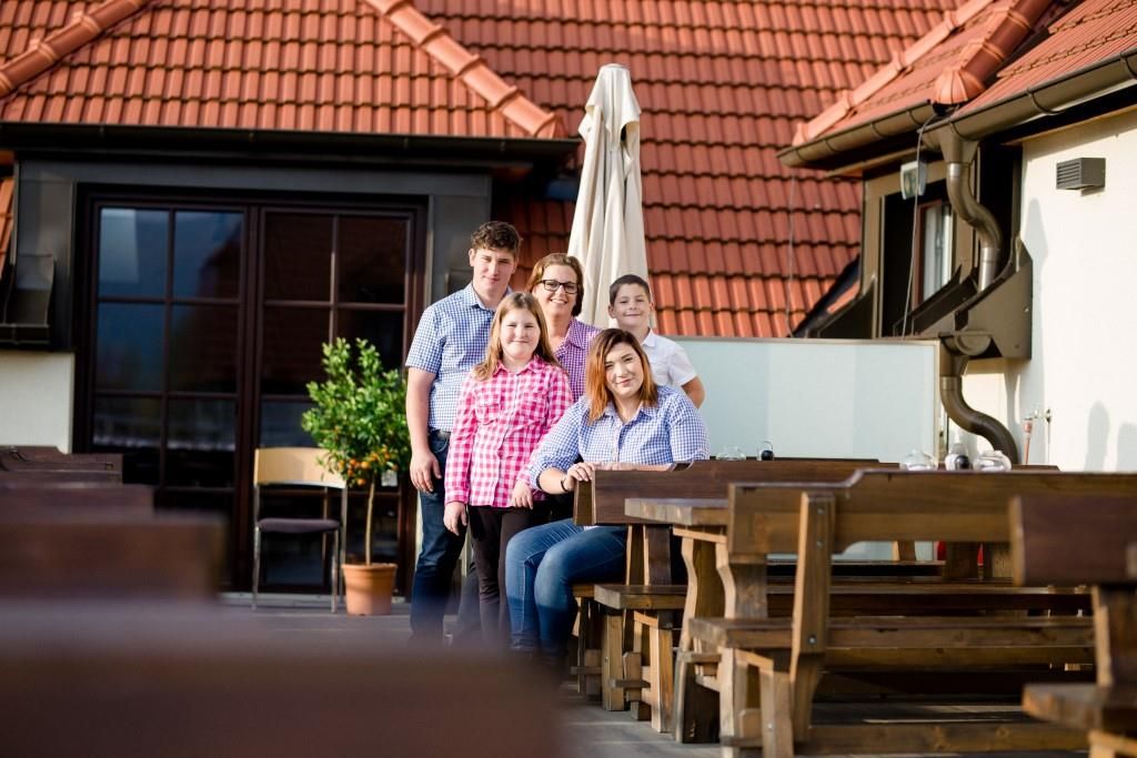 A family poses on a terrace with wooden tables and red roof tiles in the background.