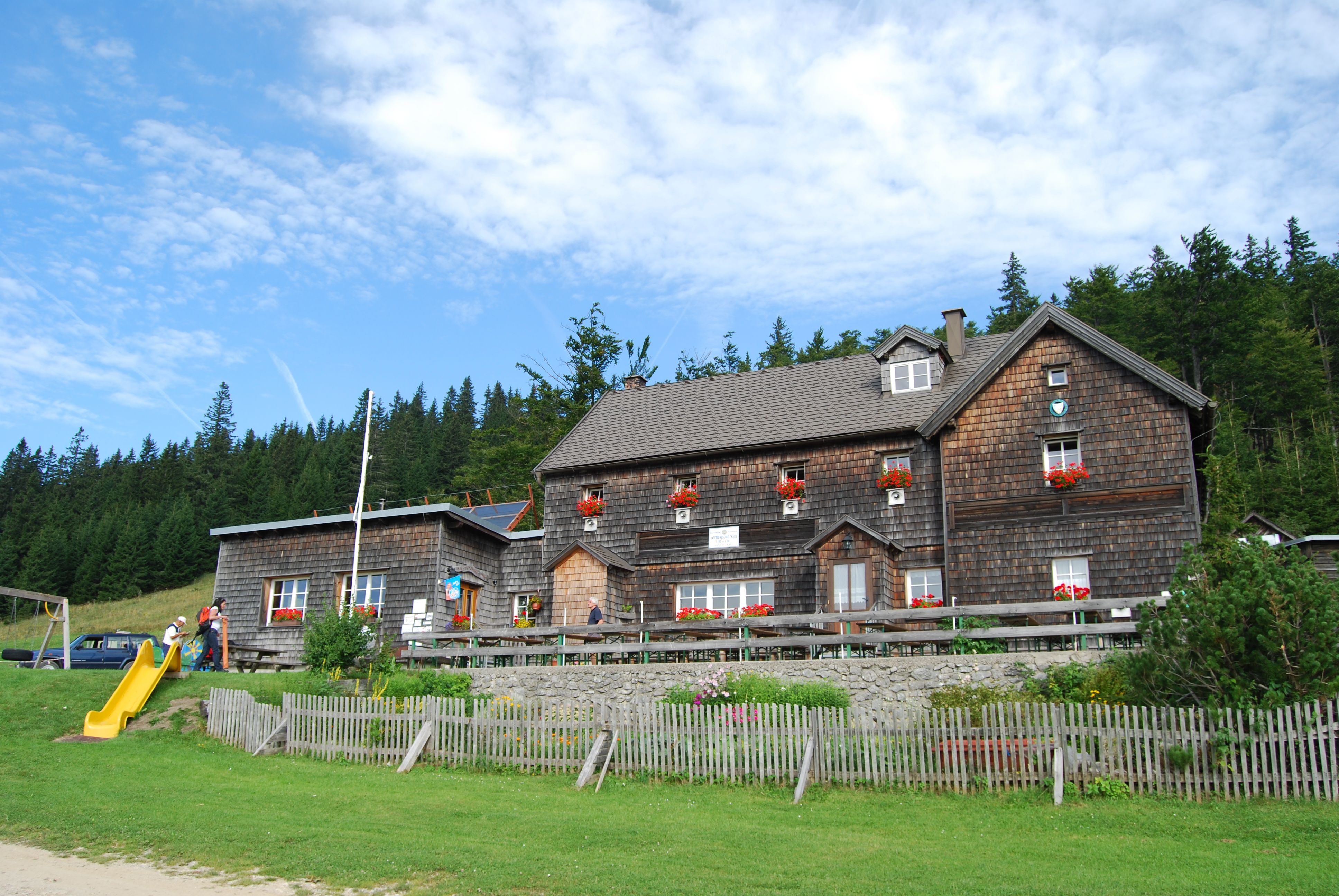 A traditional wooden house with red flower boxes in front of a forest and blue sky.