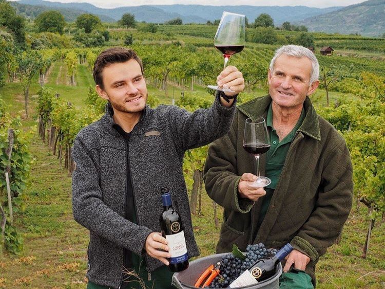 Two men in a vineyard holding glasses of red wine and smiling. One of them is holding a bottle of wine; vines and hills can be seen in the background.