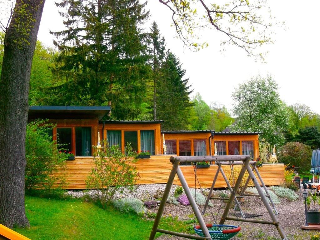 A country inn with a wooden façade and playground in the foreground, surrounded by trees.