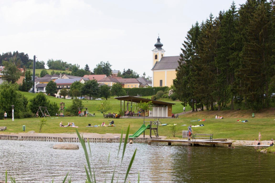 A bathing pond with sunbathing lawn, playground and a church in the background.
