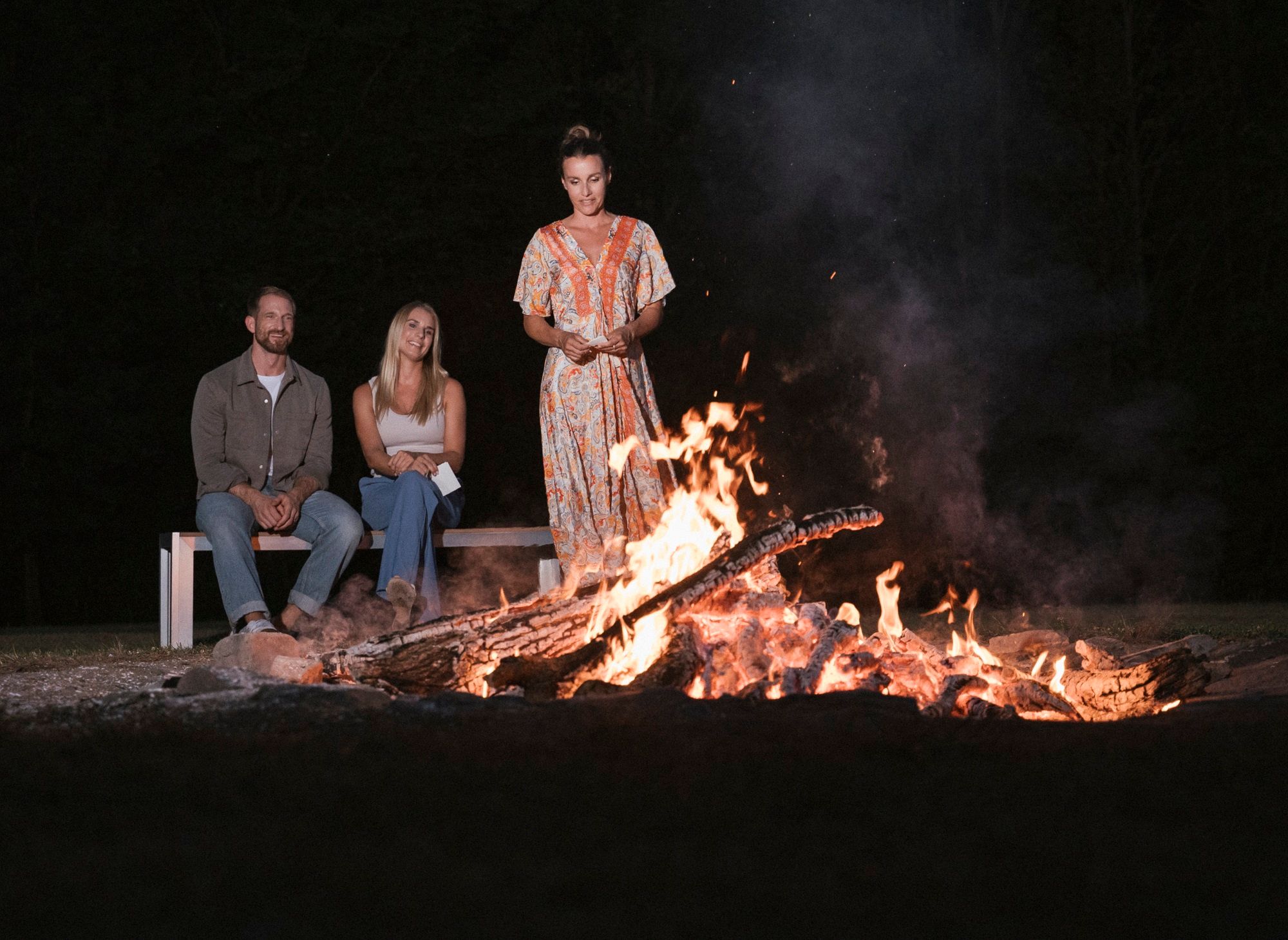Three people sit and stand around a campfire at night.