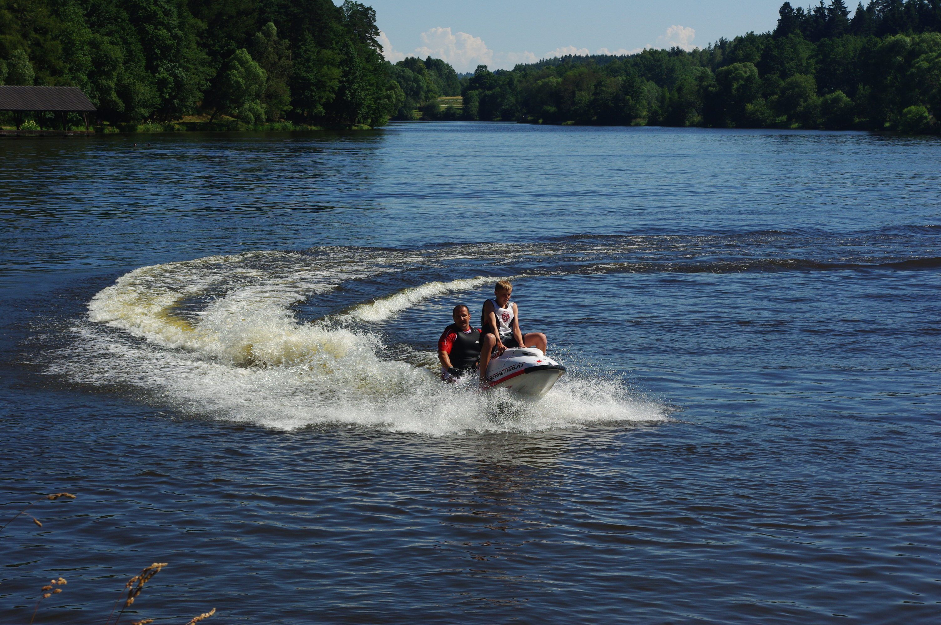 Two people ride a jet ski across a lake surrounded by trees.