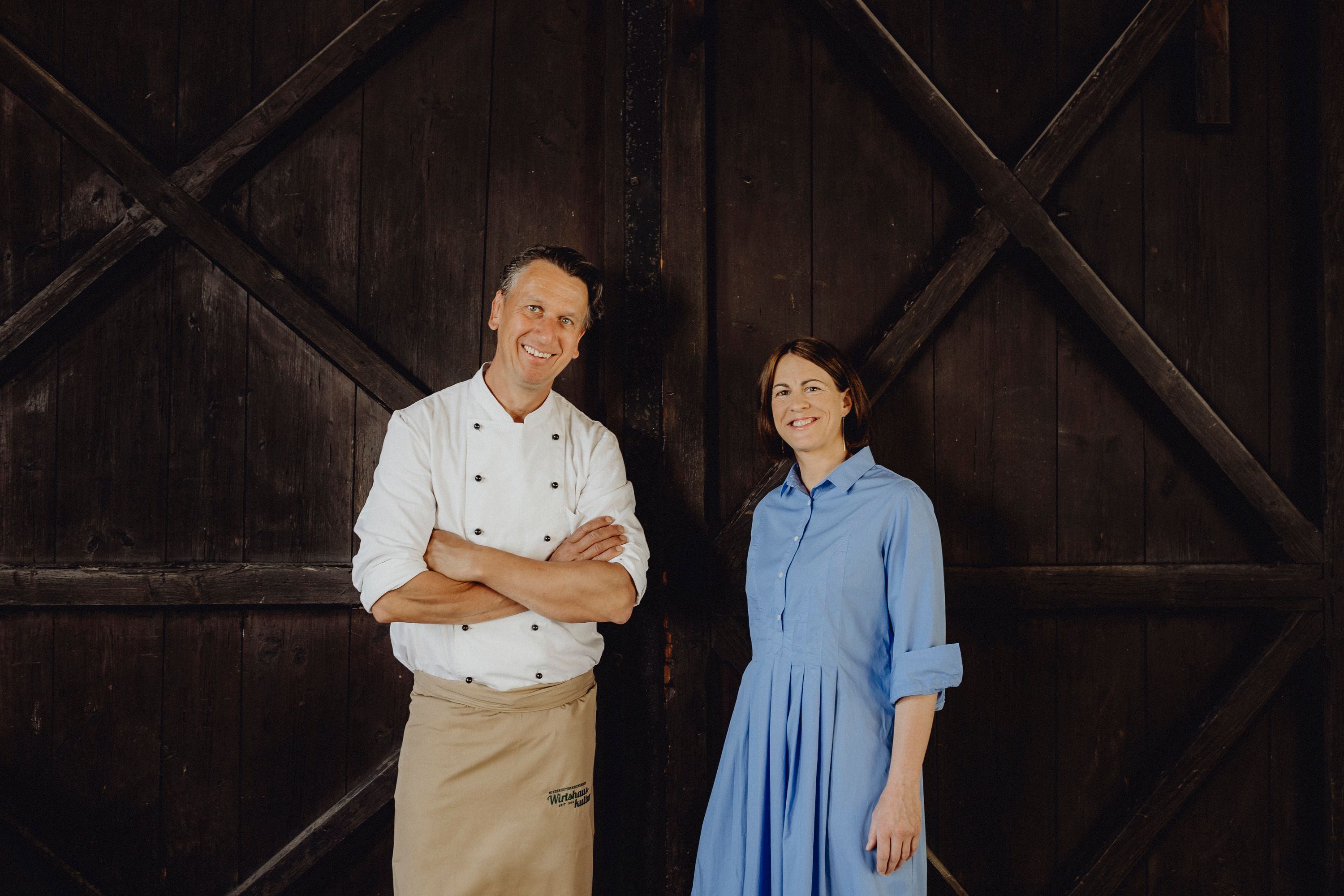 A man in chef's clothes and a woman in a blue dress stand in front of a wooden wall.