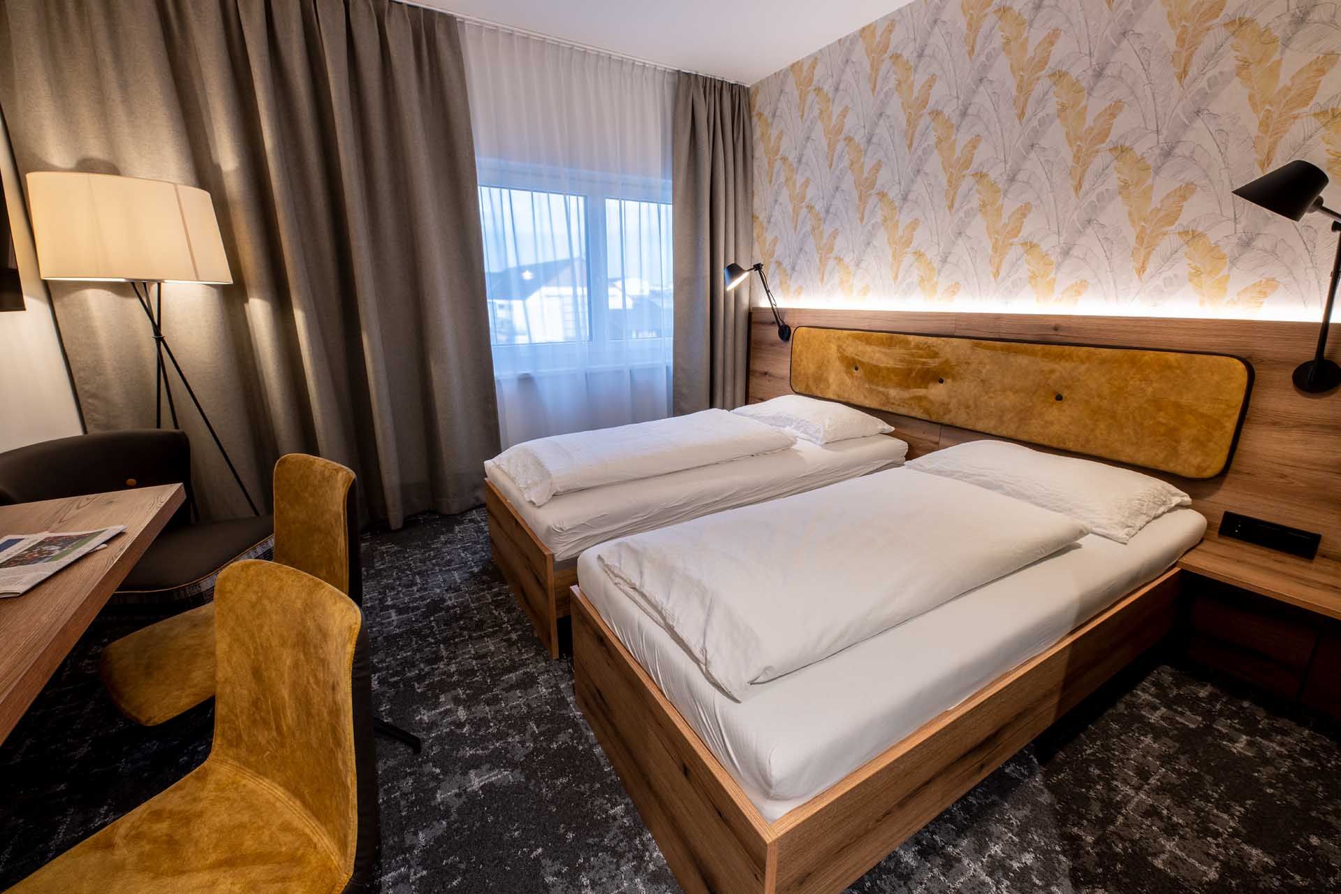 Modern guest room with two single beds, wooden furniture, yellow chairs and a floor lamp. Light-colored curtains and patterned wallpaper on the wall.