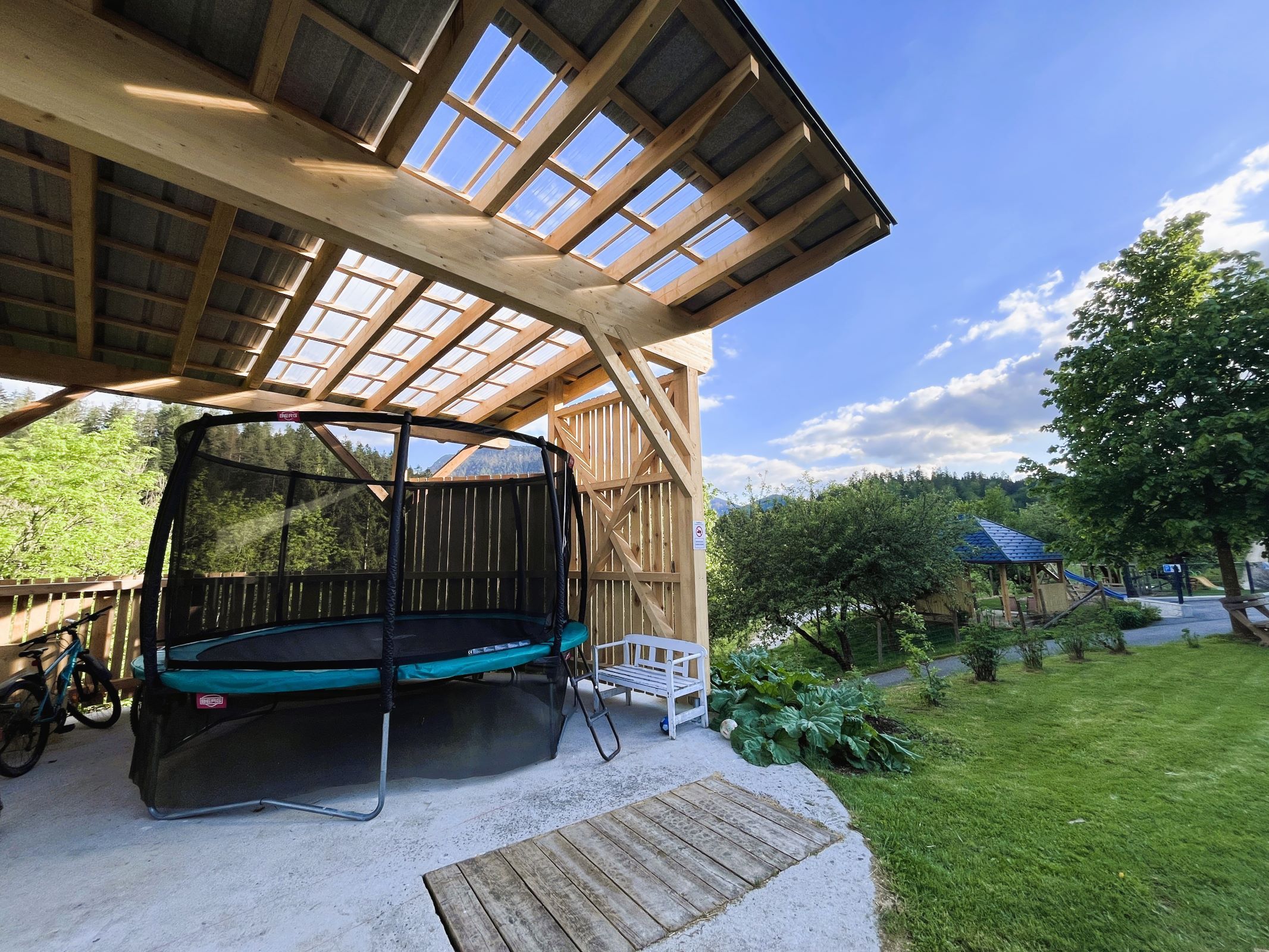 Covered trampoline in a garden with a wooden roof and surrounding nature.