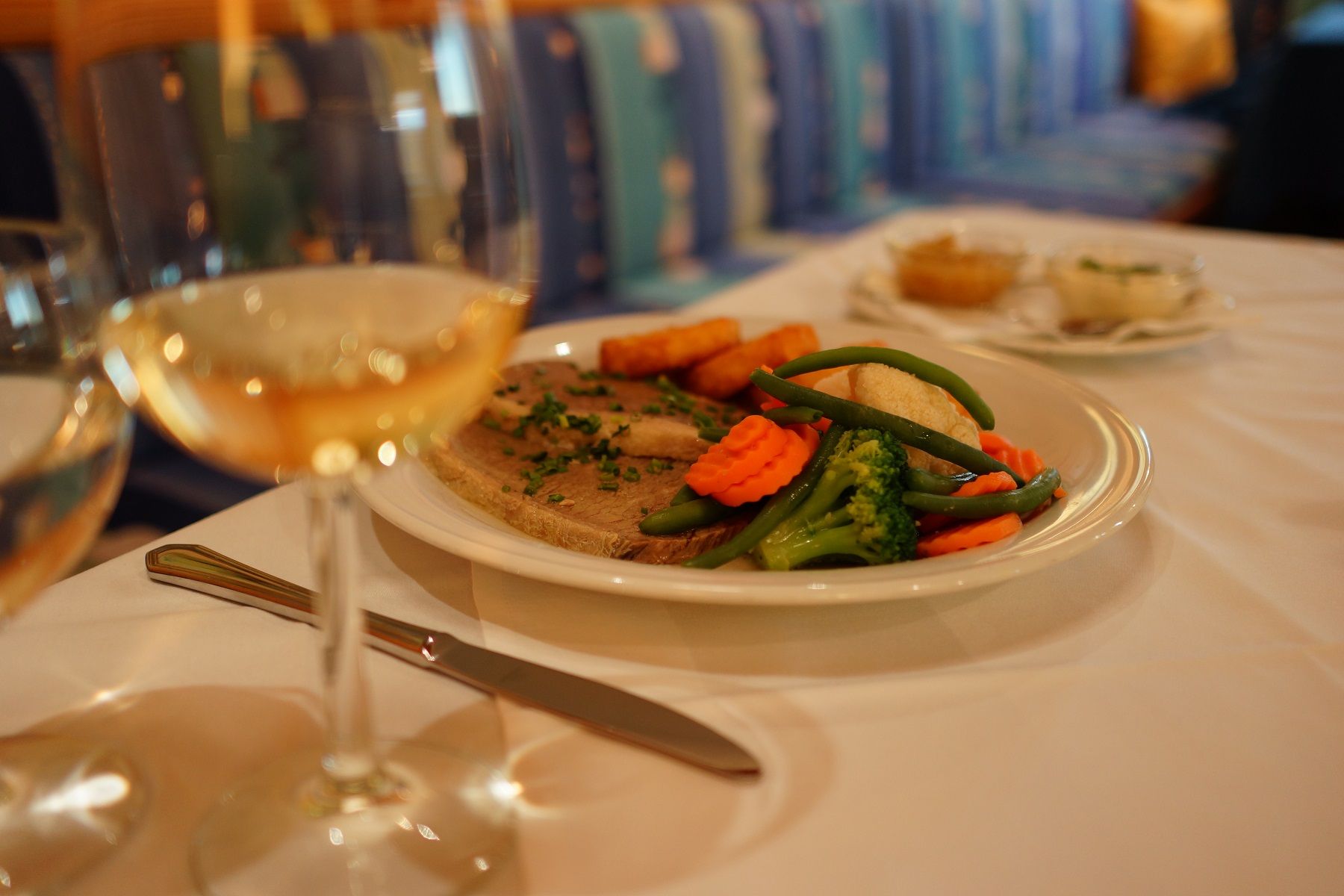 A plate of meat, vegetables and potatoes on a table, with a glass of white wine next to it.