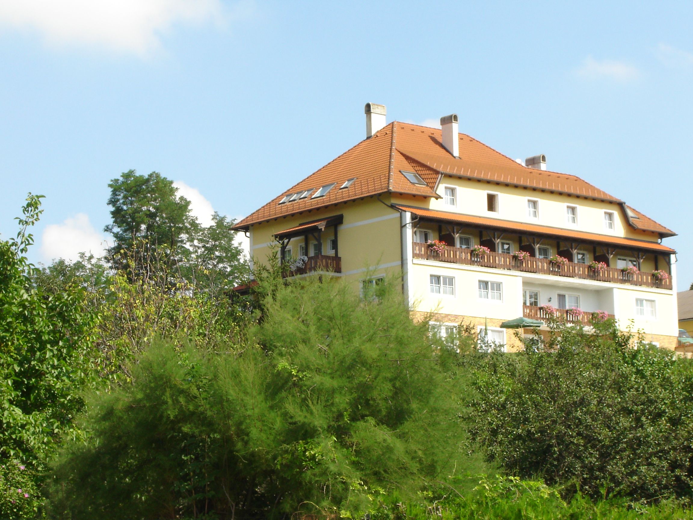 A large, multi-storey building with a red roof and balconies, surrounded by trees and plants.