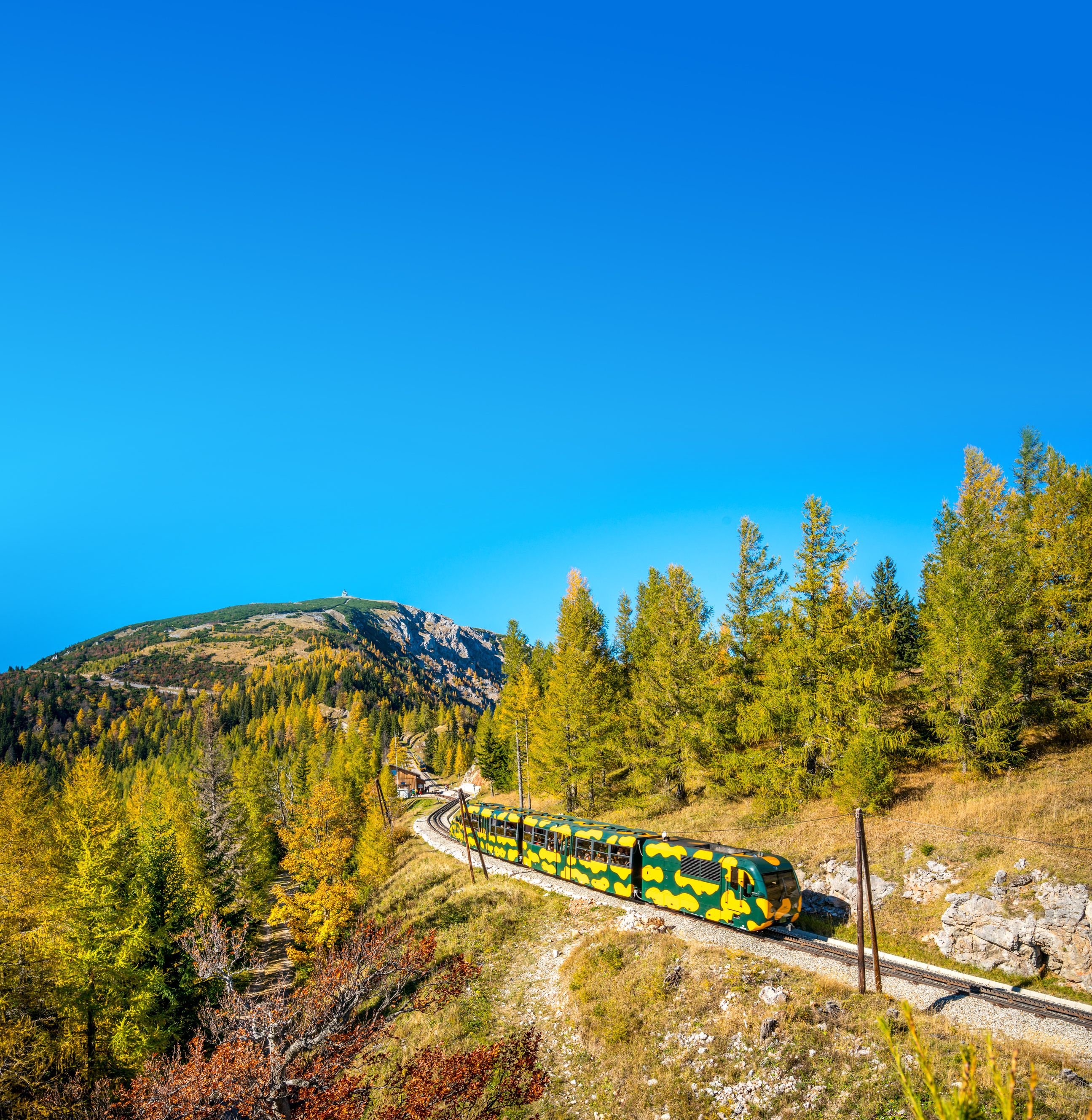A train in salamander design travels through an autumnal mountain landscape with a blue sky.