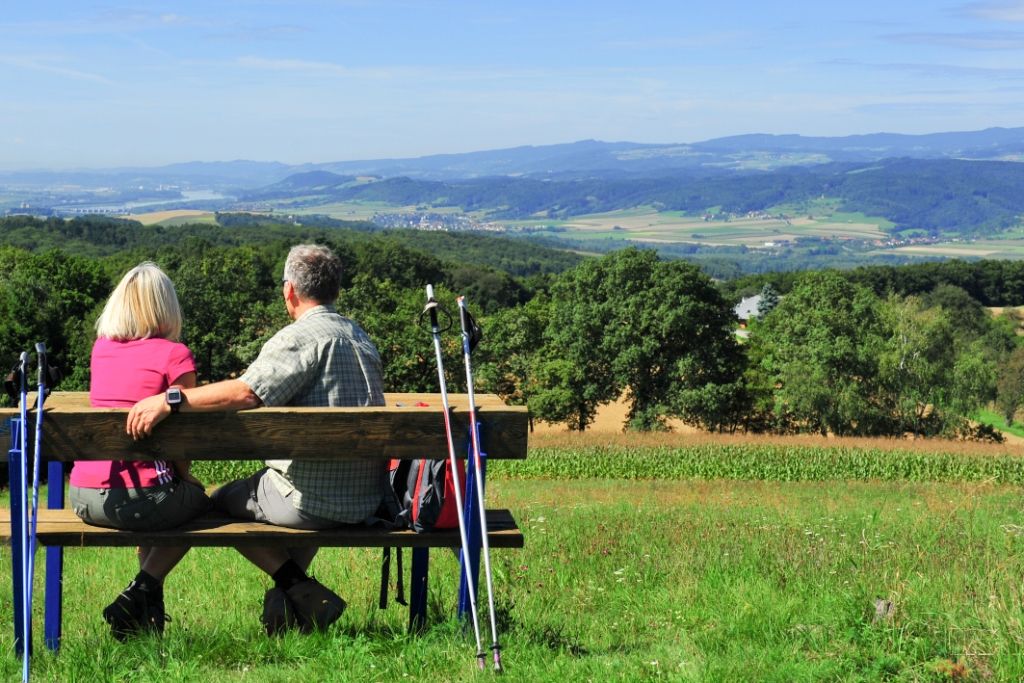 A couple sits on a bench with walking sticks and looks out over a hilly landscape.