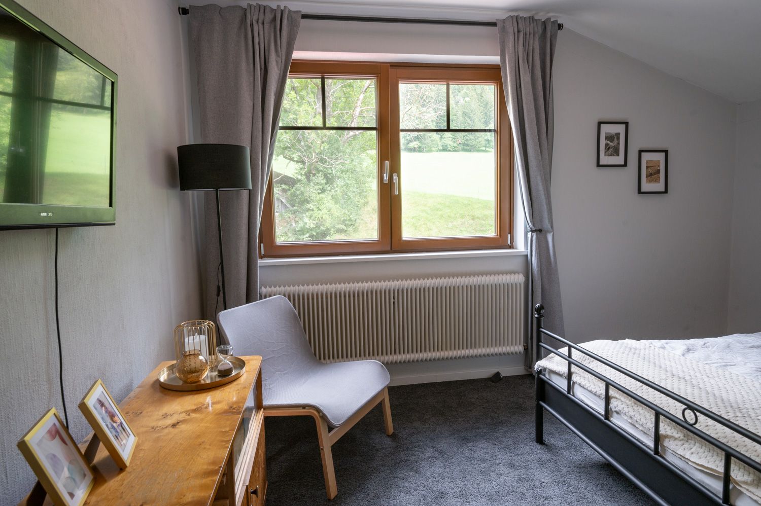 Bedroom with gray carpet, bed, armchair, TV and window with a view of the greenery.