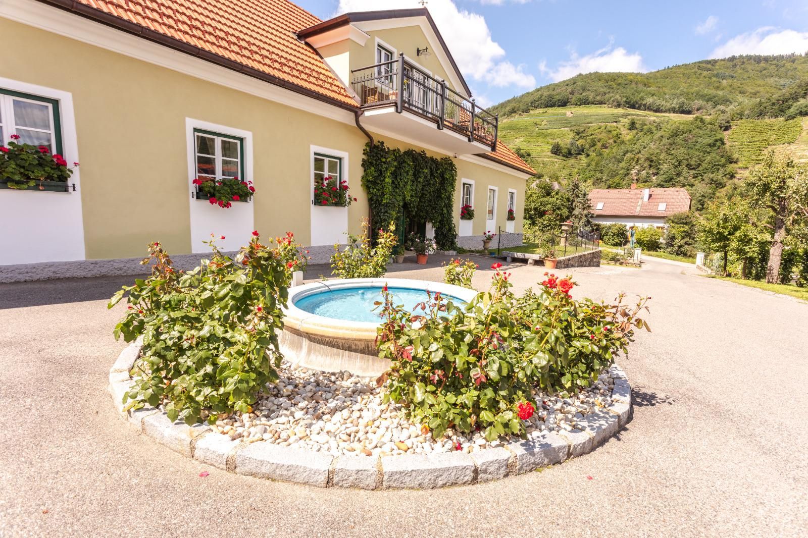 Entrance to a winery with fountain and flowers, surrounded by hills.