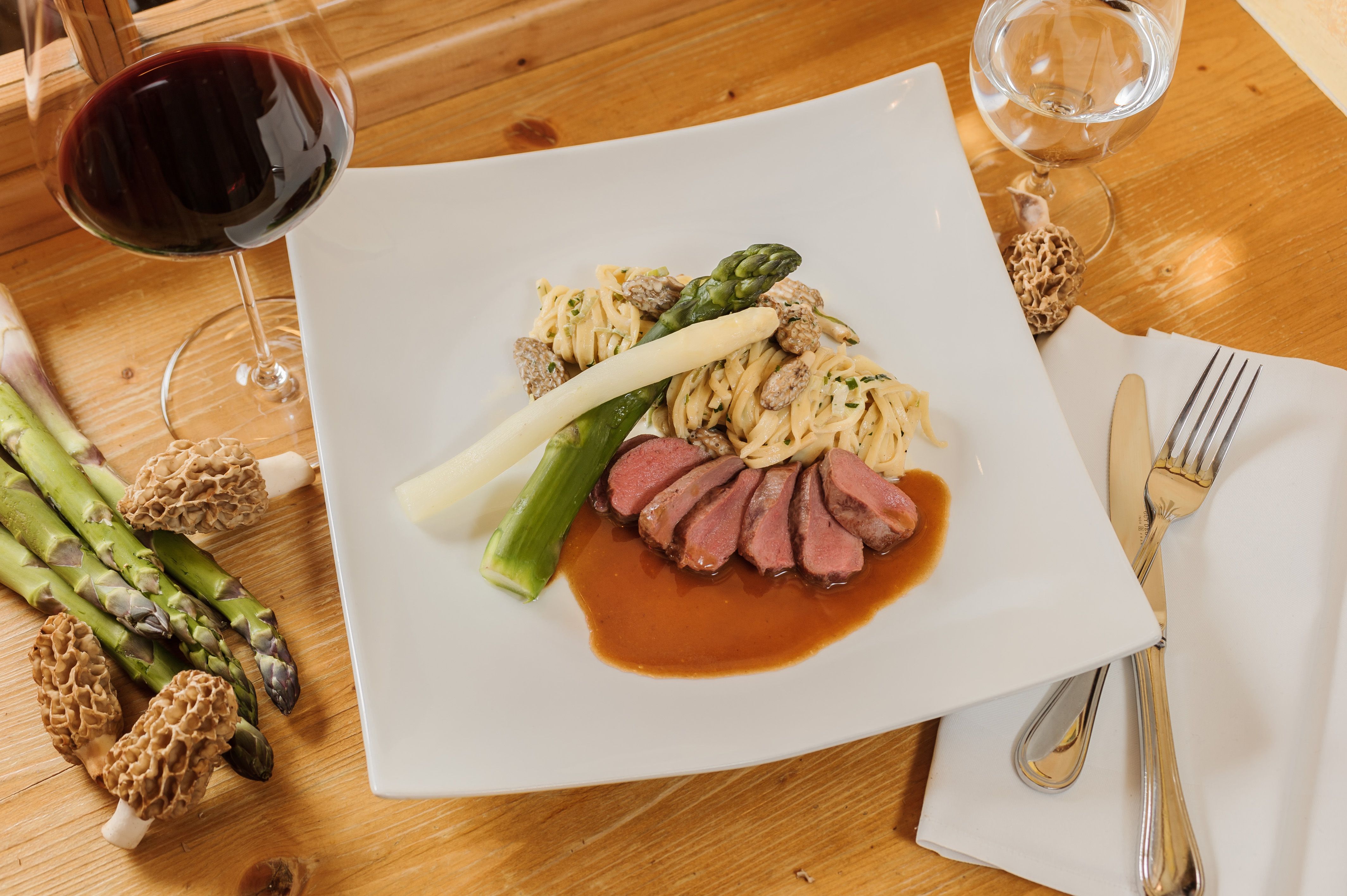 Gourmet dish with meat, asparagus, pasta and morels on a white plate, with a glass of red wine next to it.