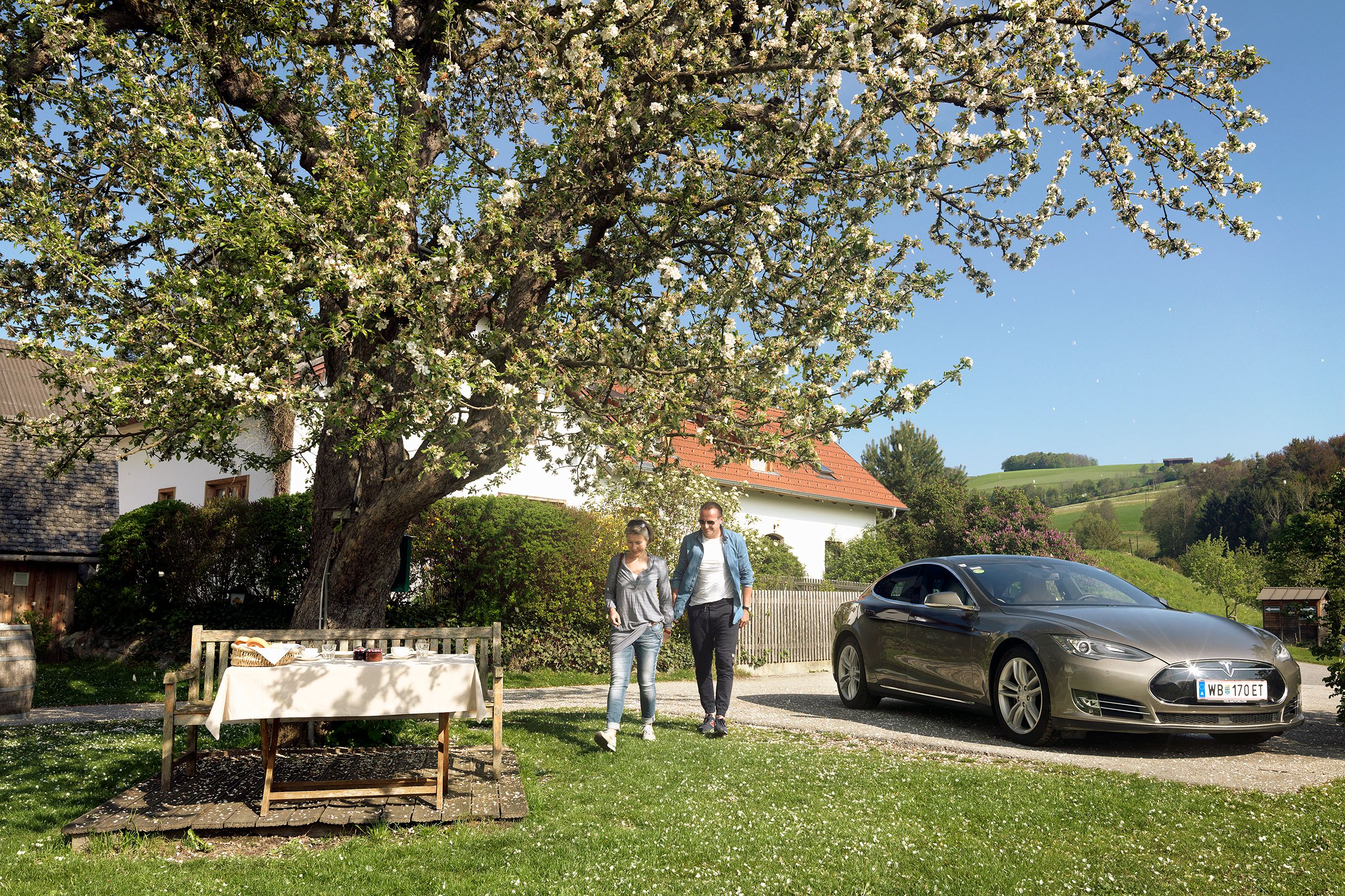 A couple walks next to a parked electric car under a blossoming tree in a rural garden.