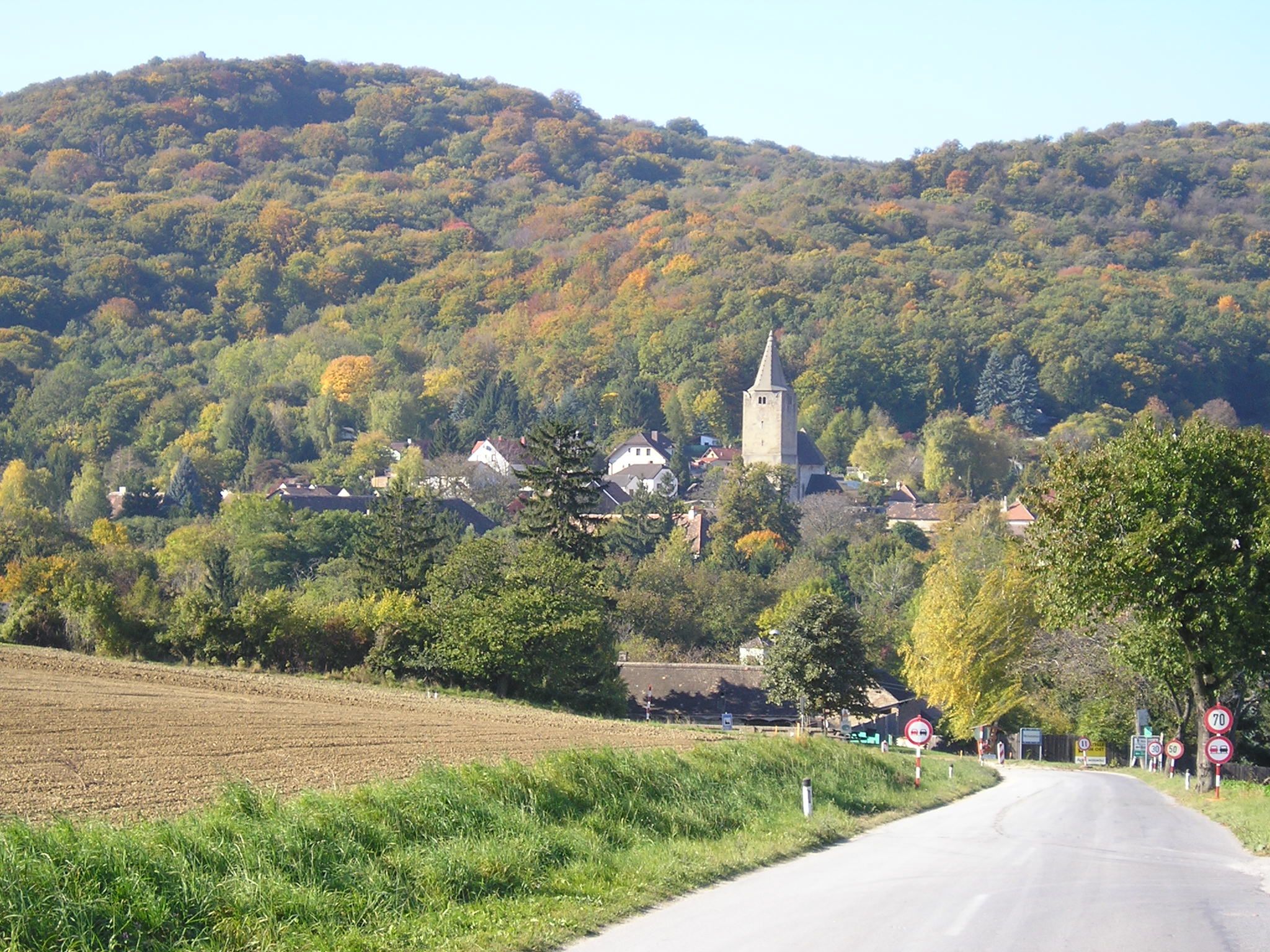A picturesque landscape with a road leading to a village with a church, surrounded by wooded hills in the fall.