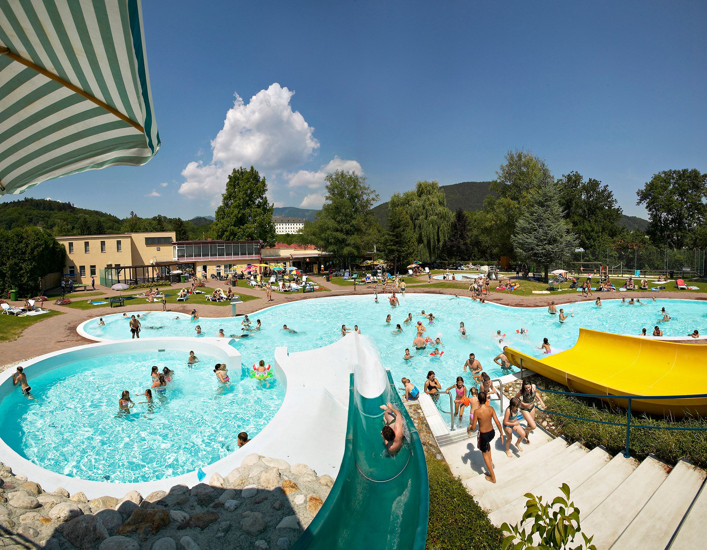 An outdoor pool with several pools, water slides and lots of people on a sunny day.