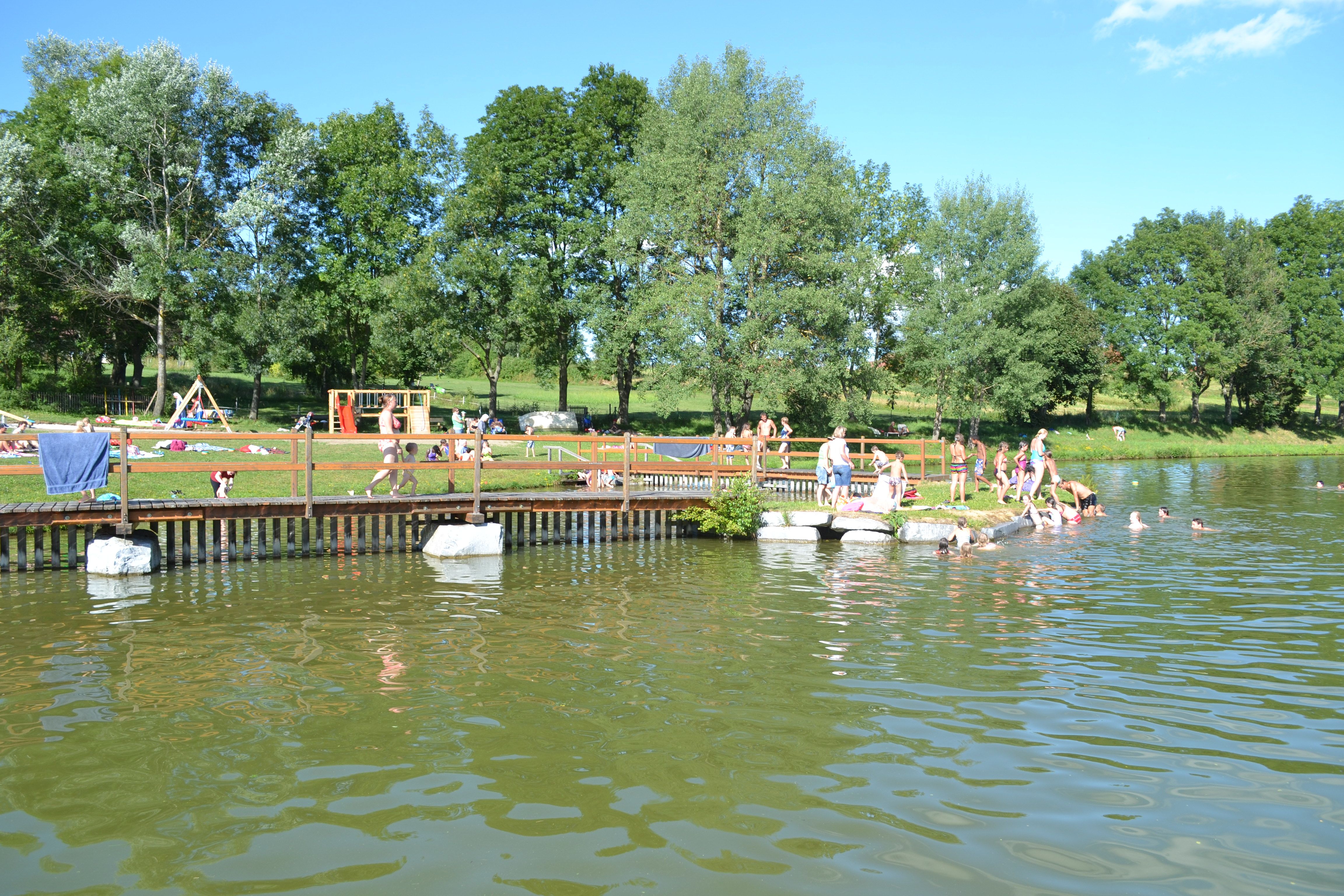 People swimming and relaxing at a bathing pond with a jetty and playground in the background.