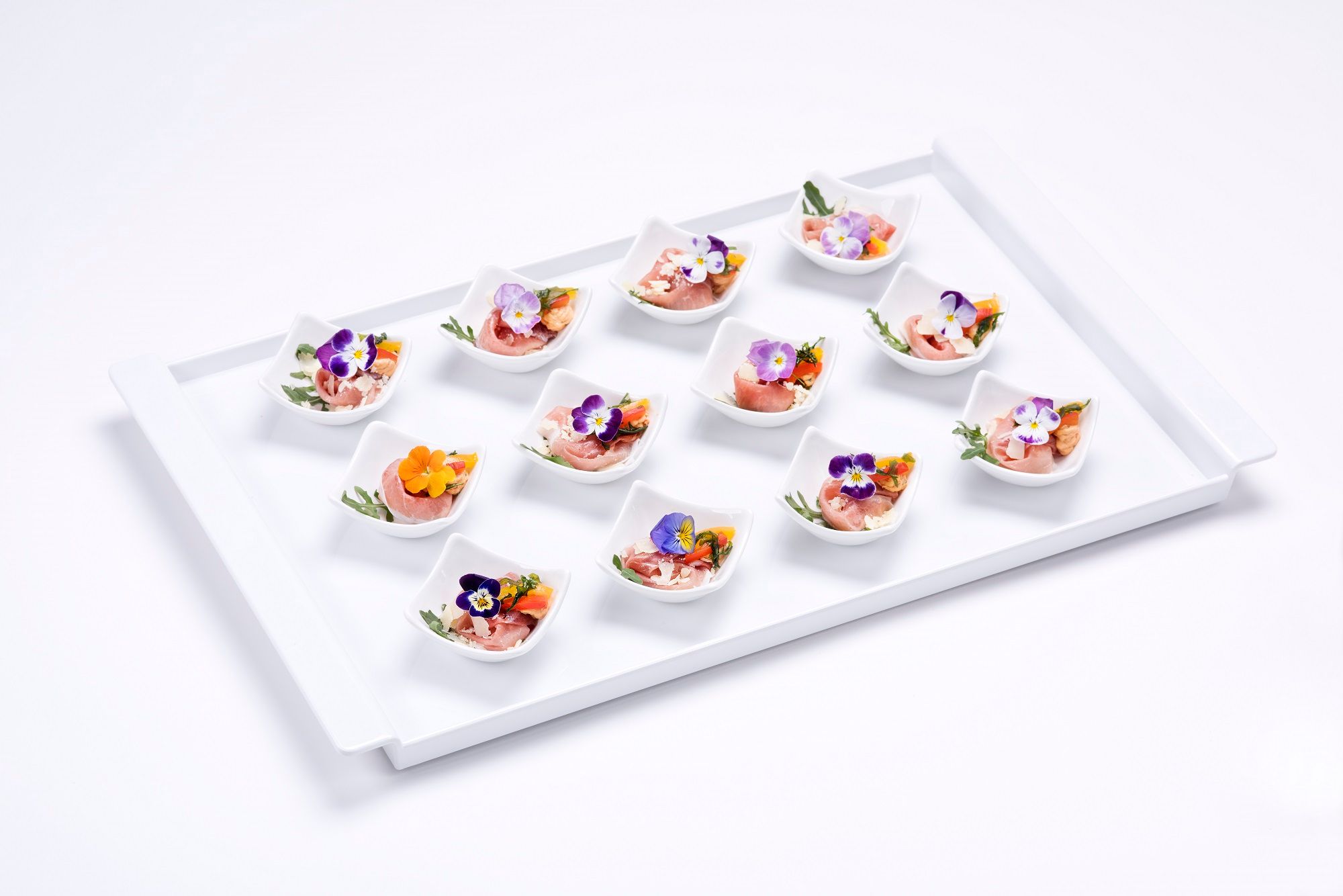 A tray with twelve small bowls decorated with ham, edible flowers and herbs.