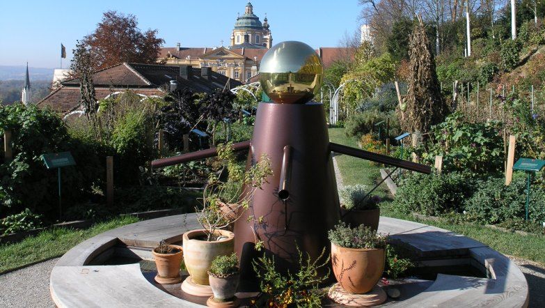 A decorative garden with a sculpture in the foreground and a historic building in the background.