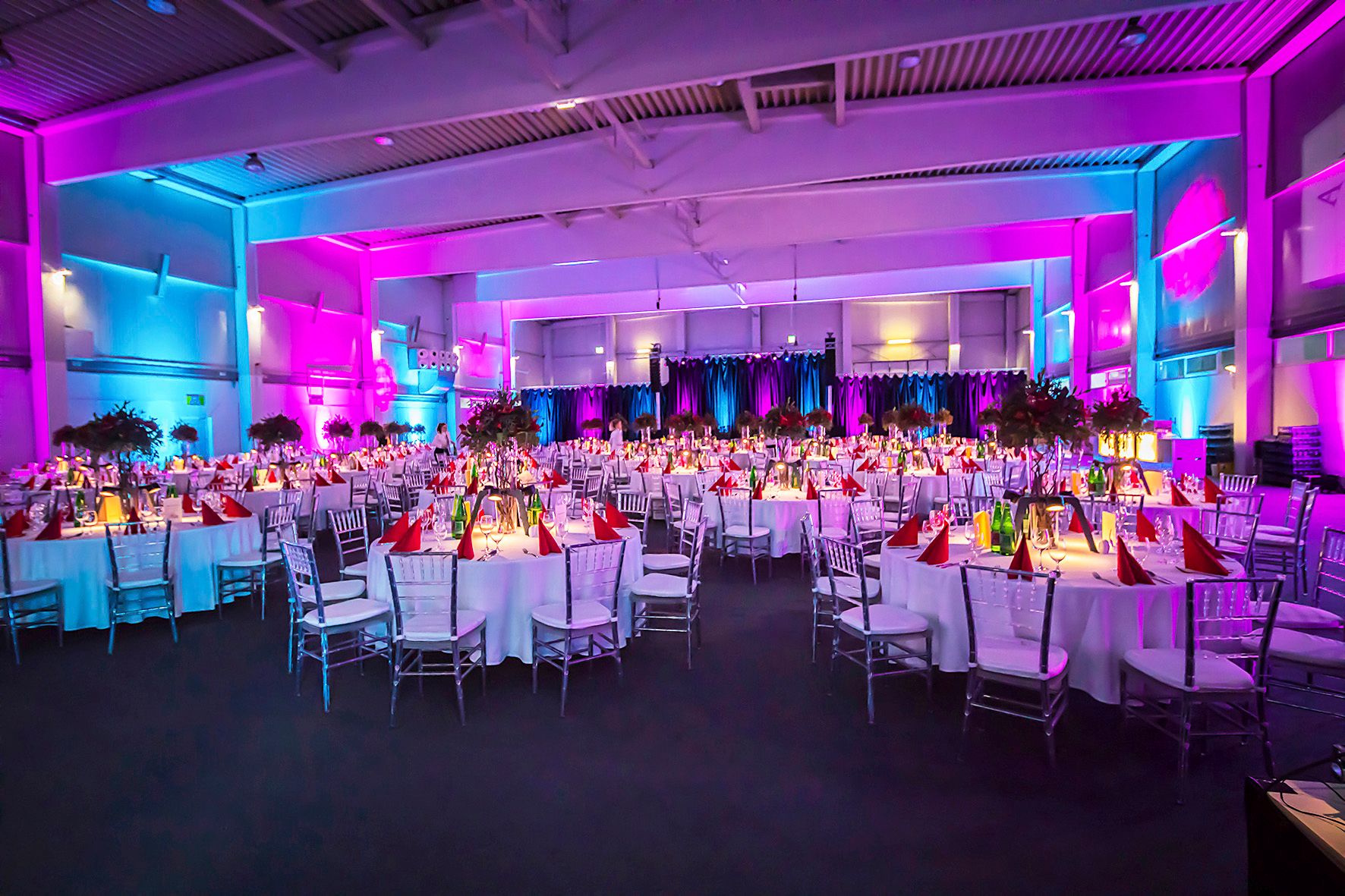 Festively decorated hall with round tables, white tablecloths and purple lighting.