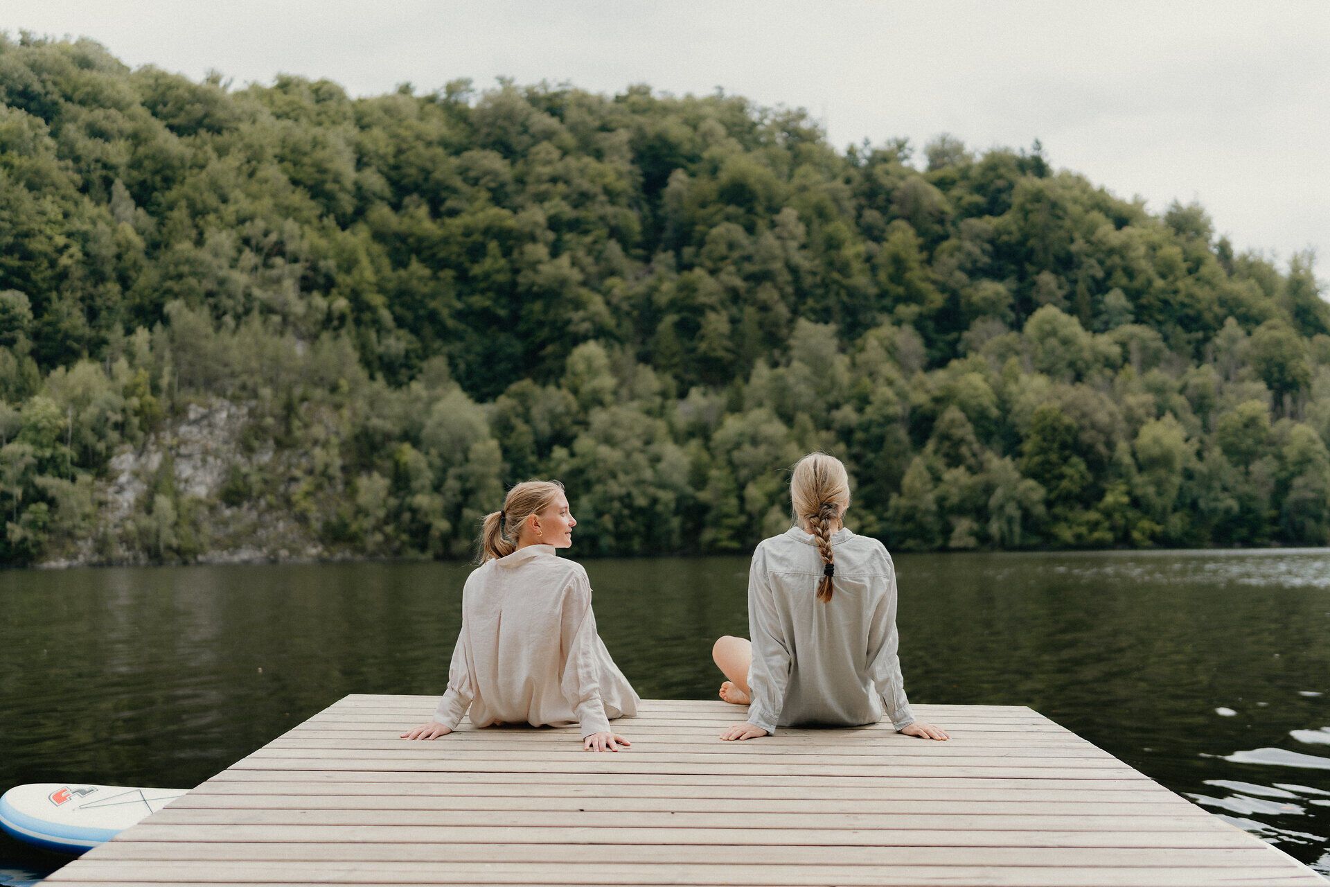 Zwei Frauen sitzen am Steg bei einem See und schauen auf die vorliegende Landschaft. Im Hintergrund sieht man den grünen Wald am gegenüberliegenden Ufer.