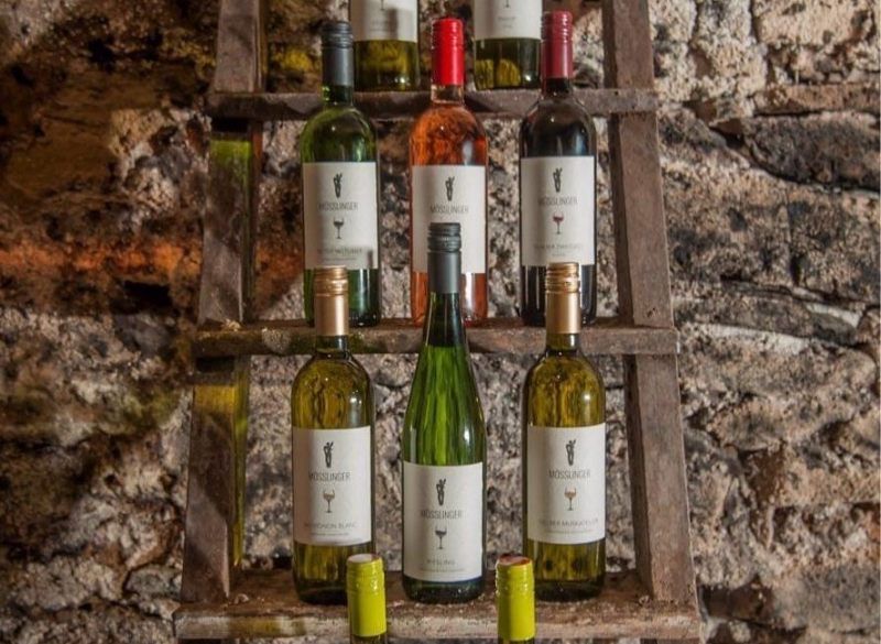 Wine bottles on a wooden shelf in front of a stone wall.
