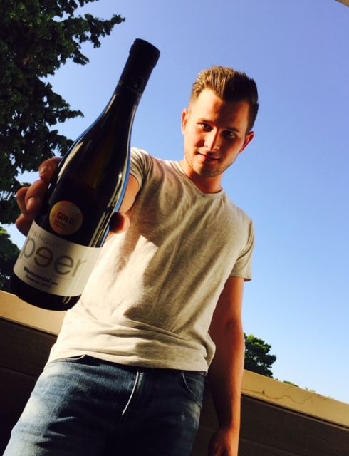 A man holds a bottle of wine up to the camera, blue sky and trees in the background.