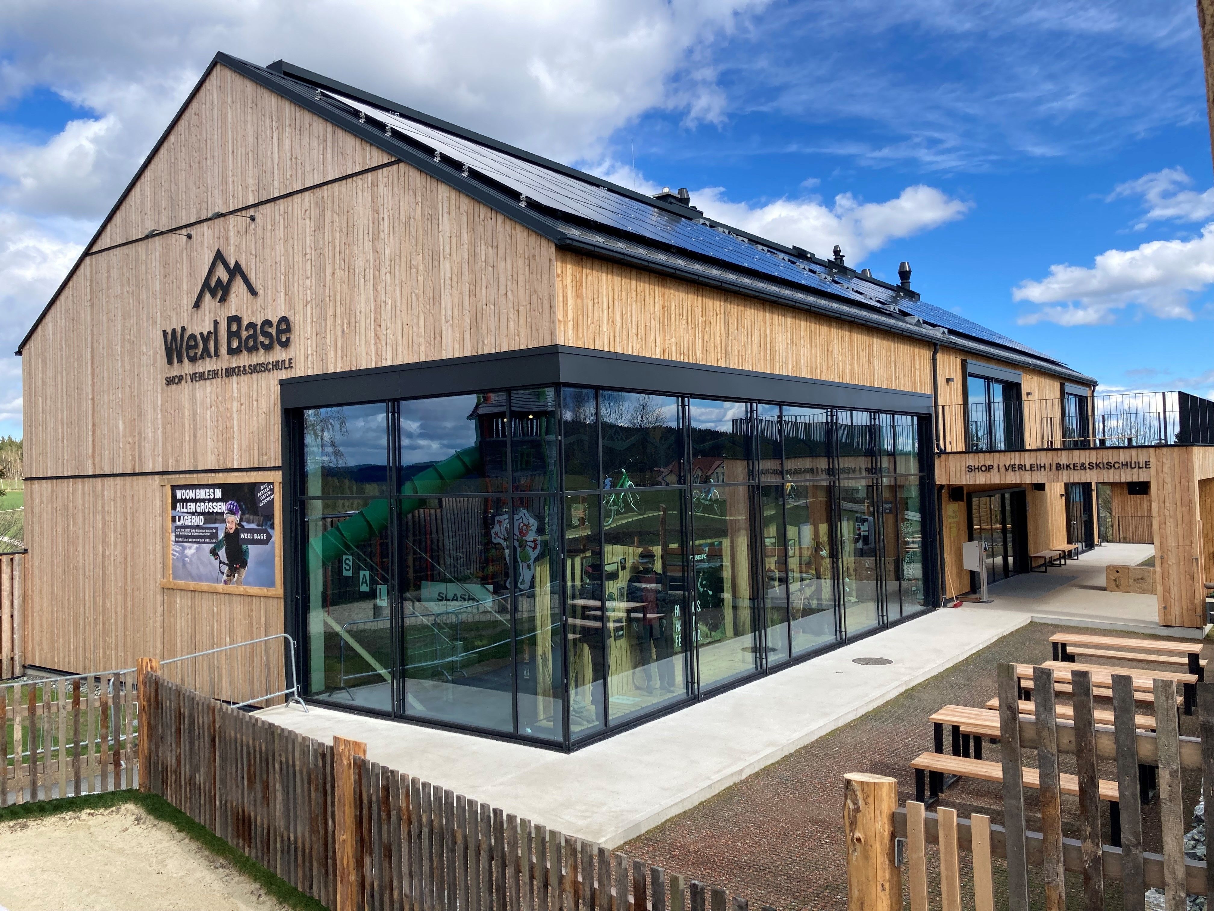 Modern Wexl Base building with wooden cladding and large glass fronts, surrounded by a fence and benches.