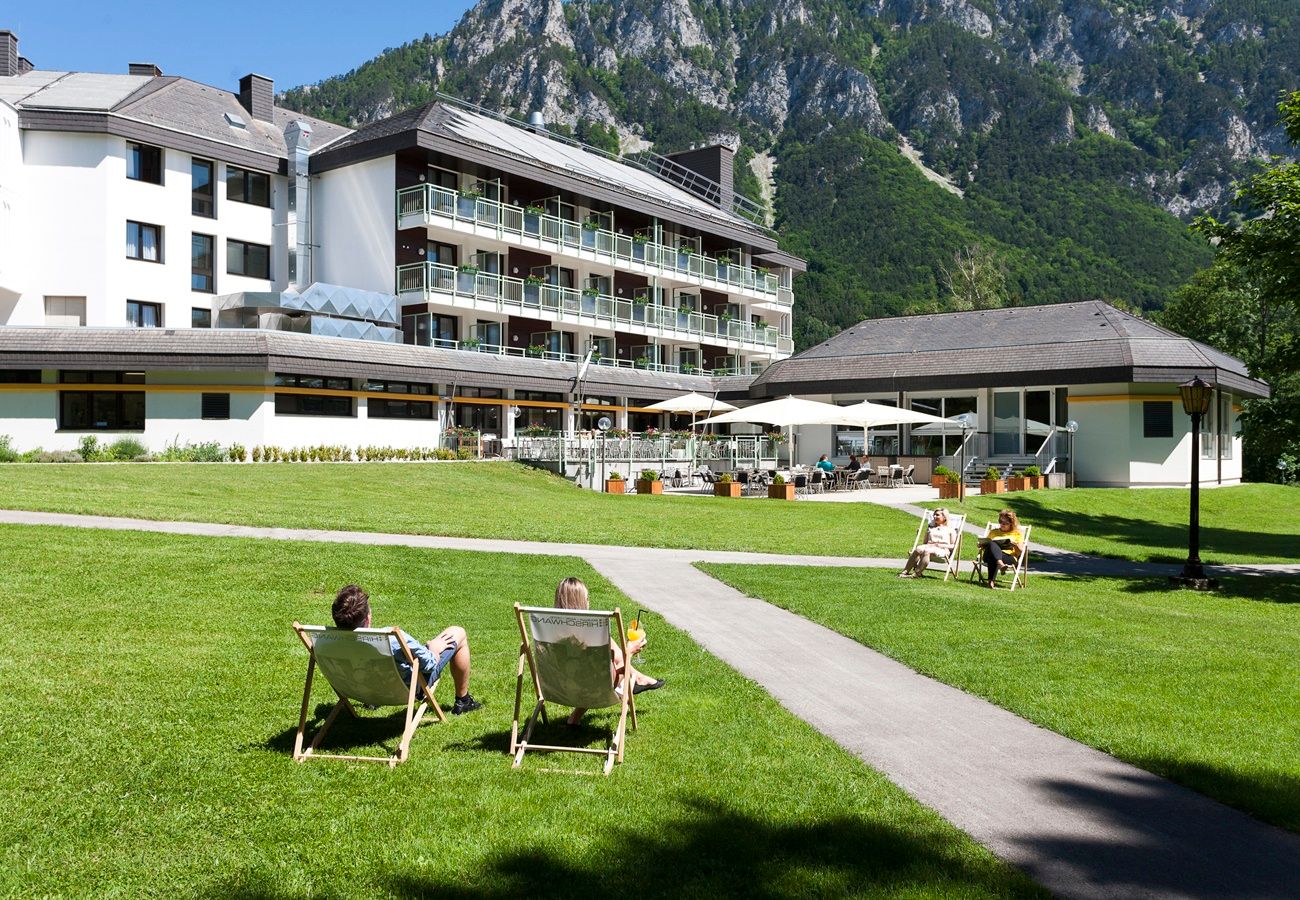 A hotel with a terrace and sun loungers on a meadow surrounded by mountains.
