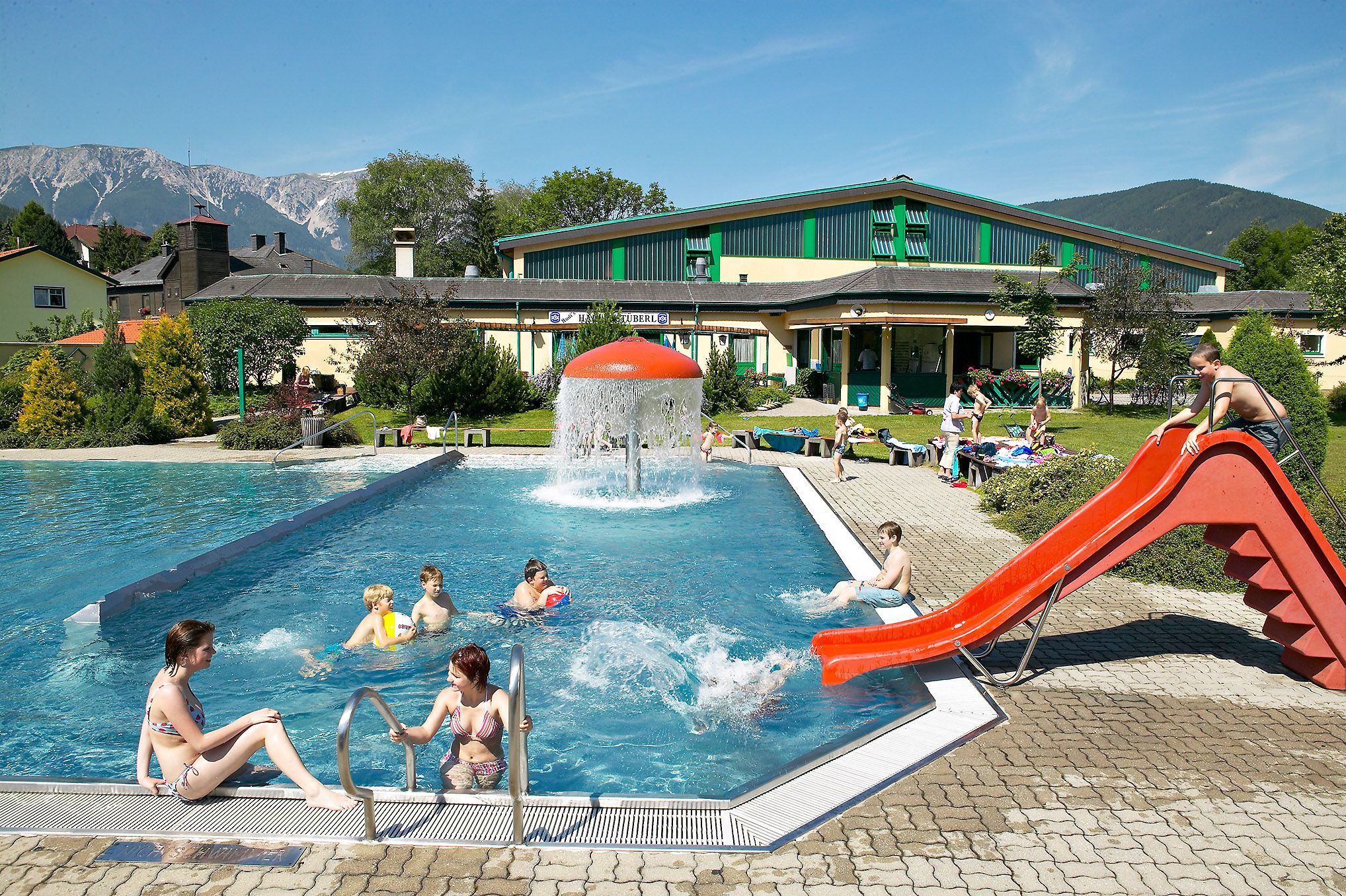 Outdoor pool with slide and mushroom fountain, surrounded by mountains.