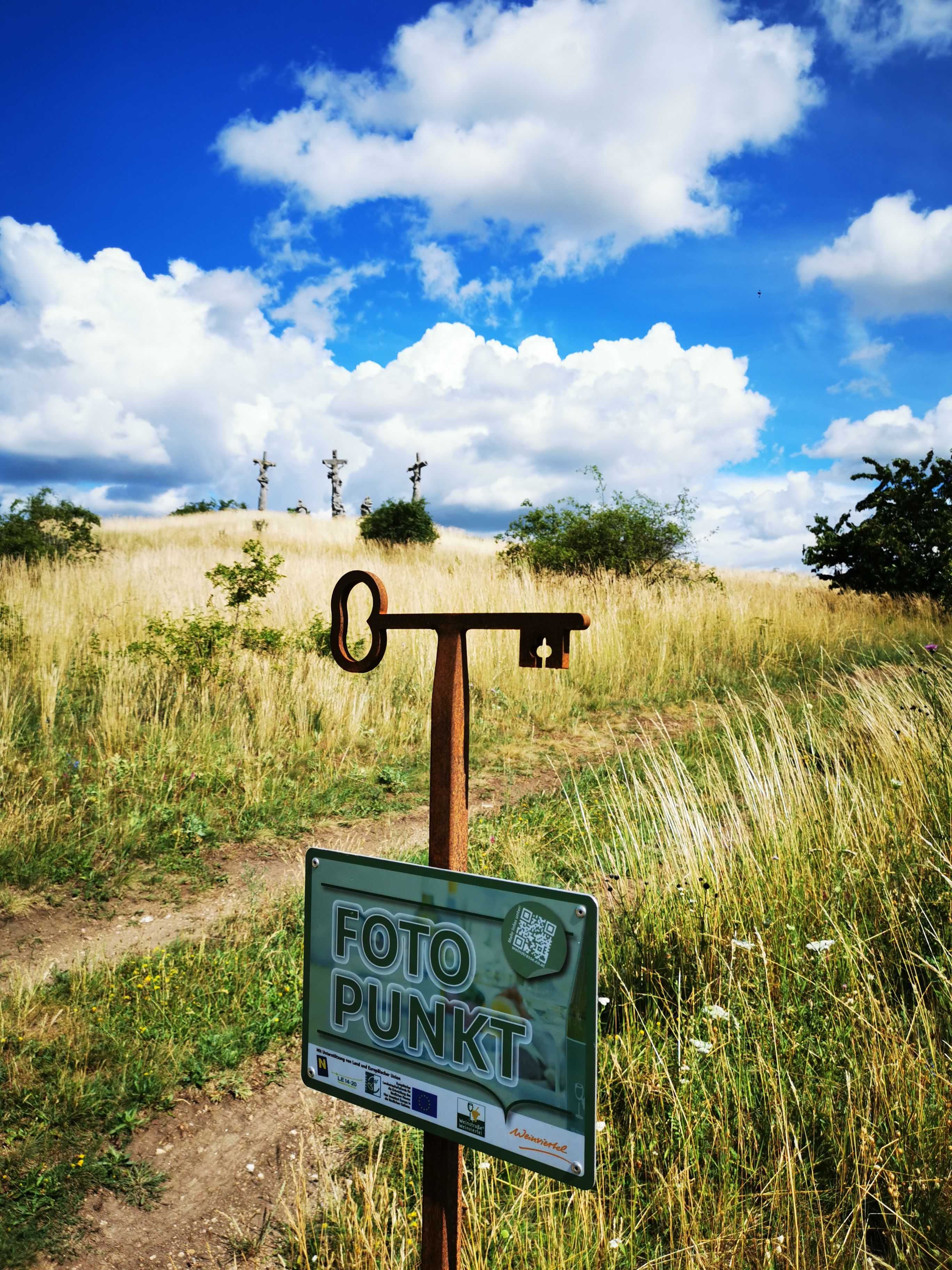 A photo point sign in front of a meadow with three crosses in the background under a blue sky with clouds.