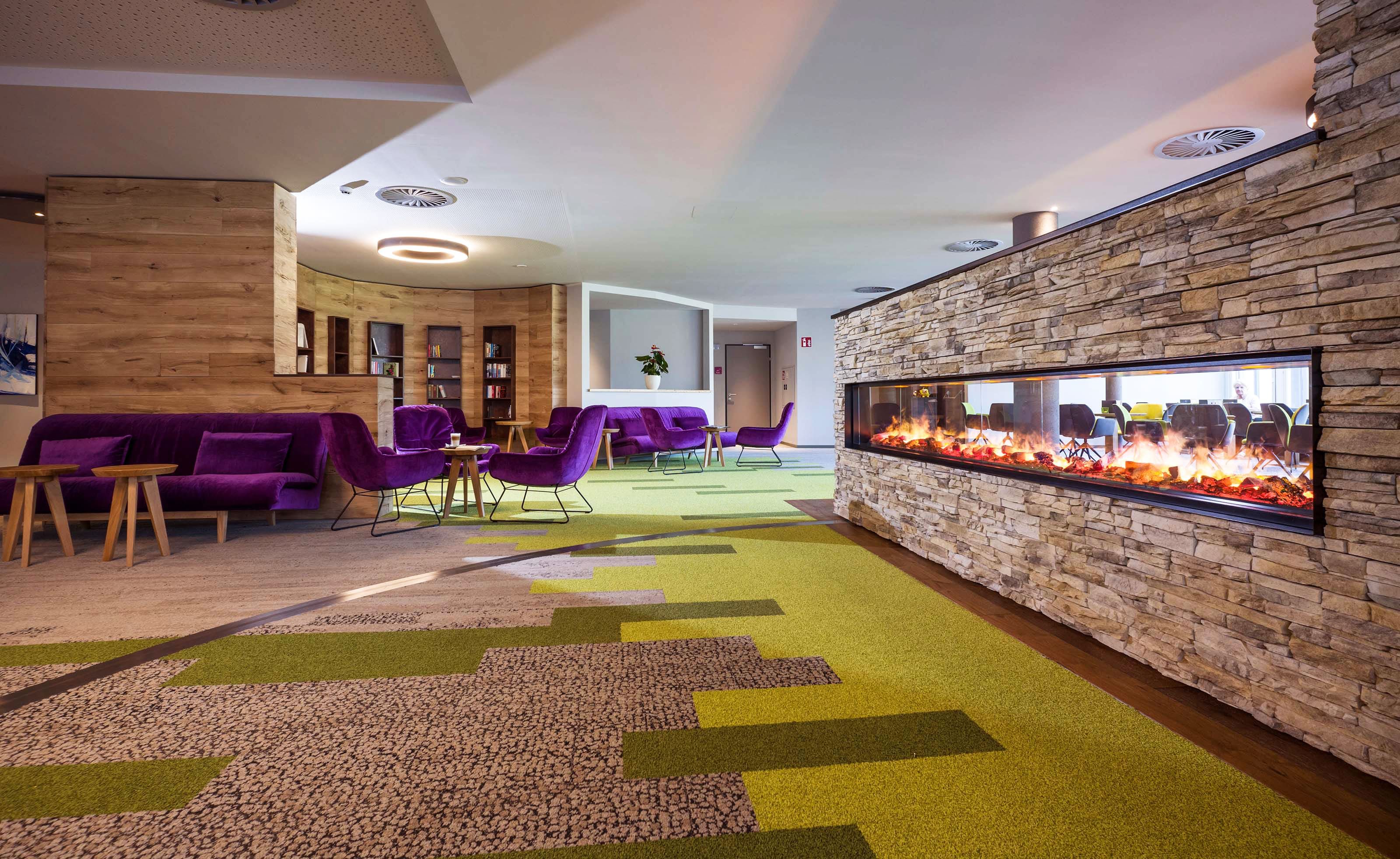 Modern hotel lobby with green-brown carpeting, fireplace, purple sofas and chairs and wooden tables.