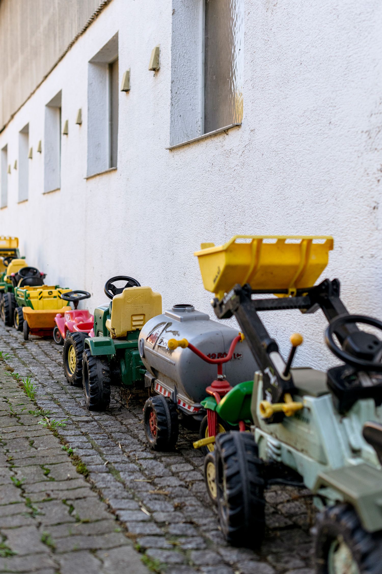 Row of toy tractors on a white wall on cobblestones.