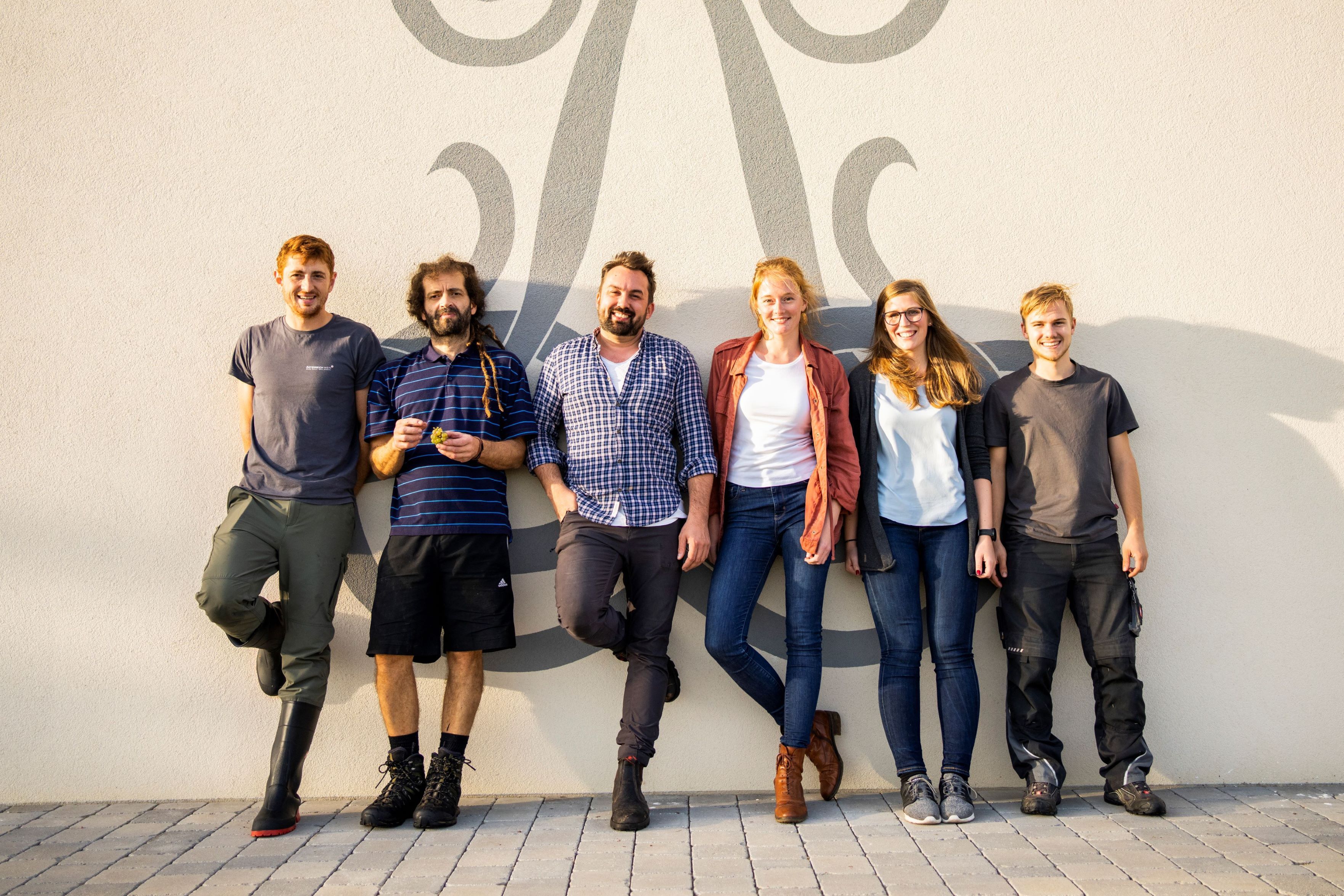 Six people stand smiling in front of a wall with a decorative pattern.