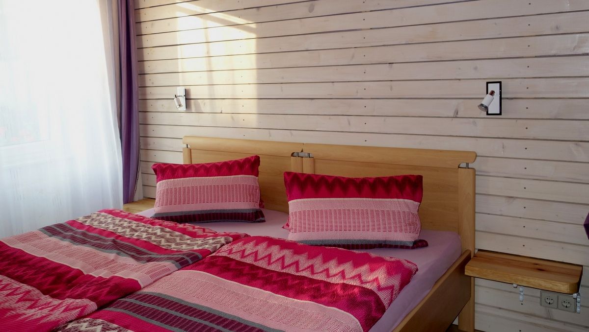 A cozy bedroom with a double bed covered with red-patterned comforters and pillows. The wall is made of light-colored wood.